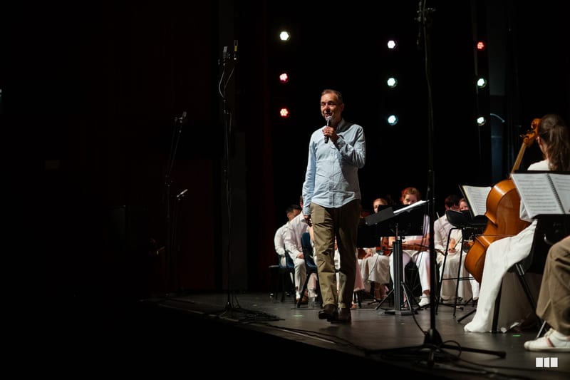 A speaker addresses the audience while an orchestra with string players performs on stage during an evening concert.