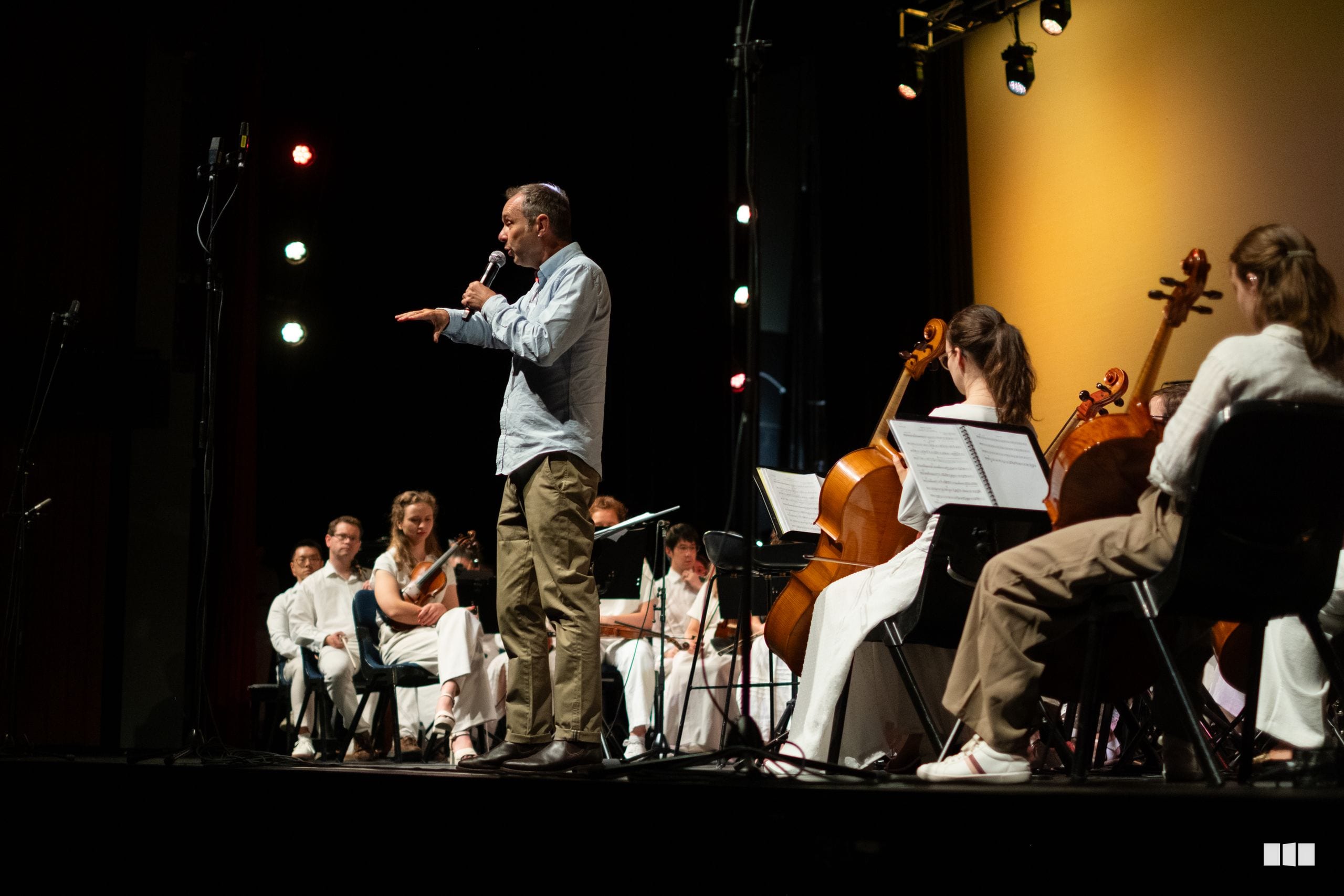 A conductor addresses an orchestra section during a rehearsal or performance, with string players visible in the ensemble.