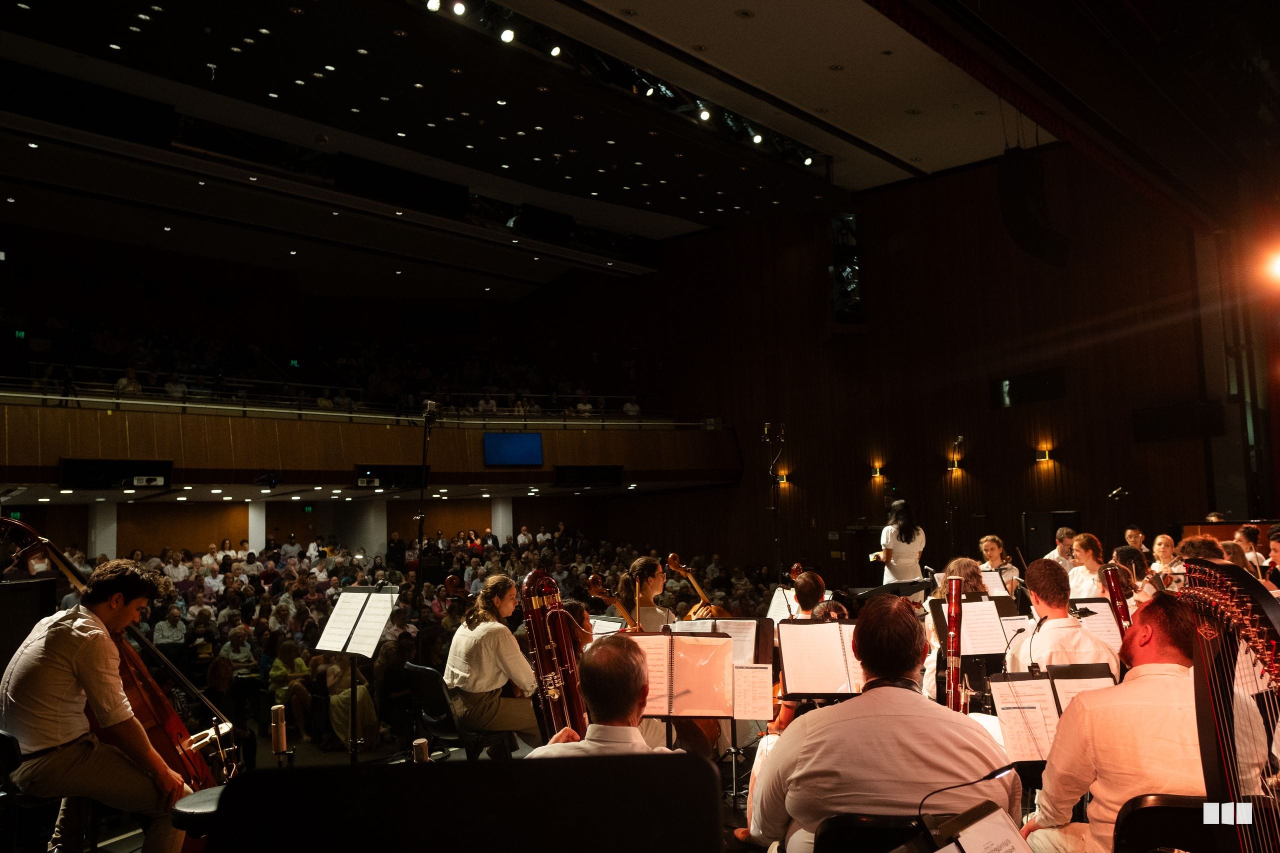 Orchestra musicians perform during a concert with a full audience visible in the auditorium seating area.
