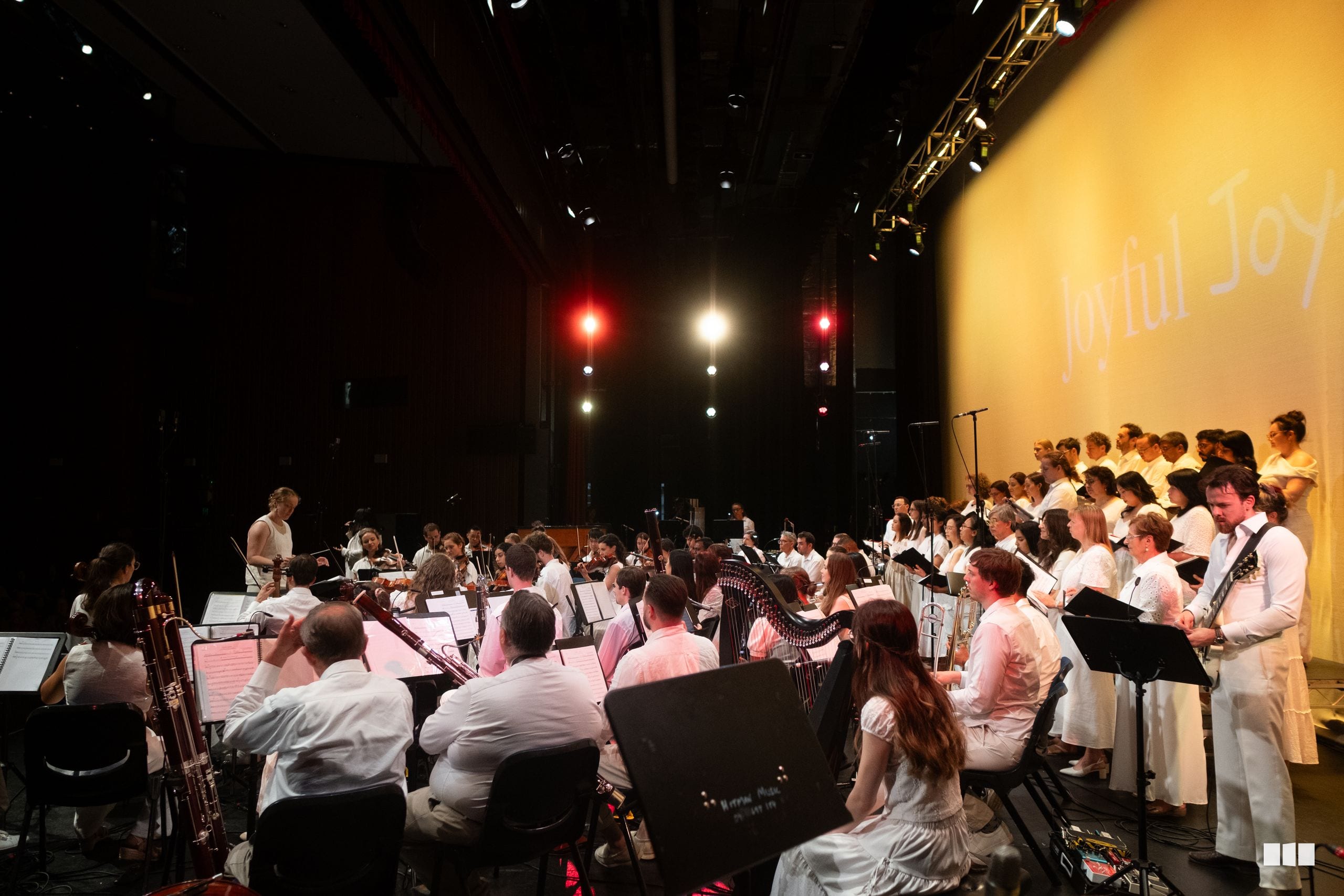 A conductor leads a combined orchestra and choir performance on stage with professional lighting and projection setup.