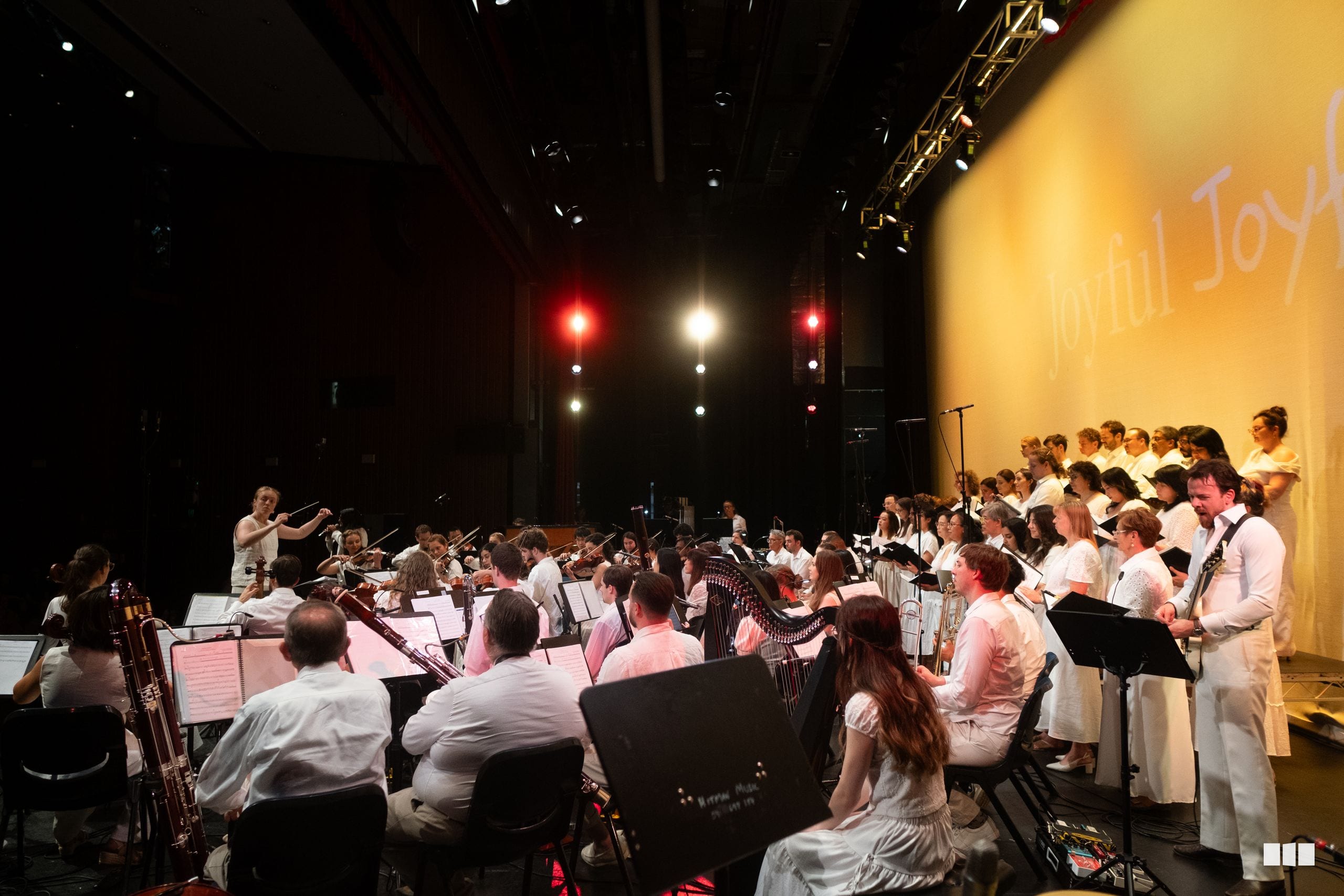 A conductor leads a combined orchestra and choir in white attire during a performance on stage with professional lighting.