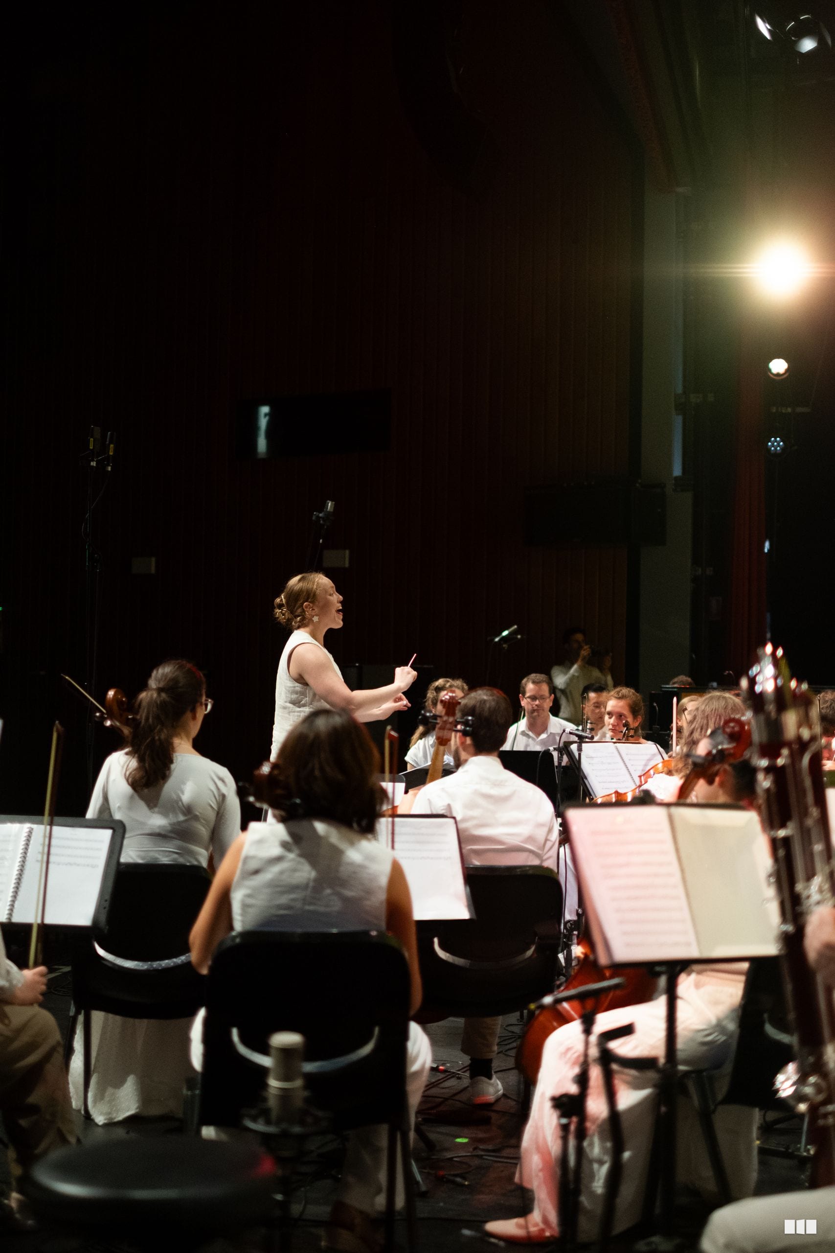 A conductor leads an orchestra during a rehearsal or performance on stage with music stands and ensemble members seated with their instruments.