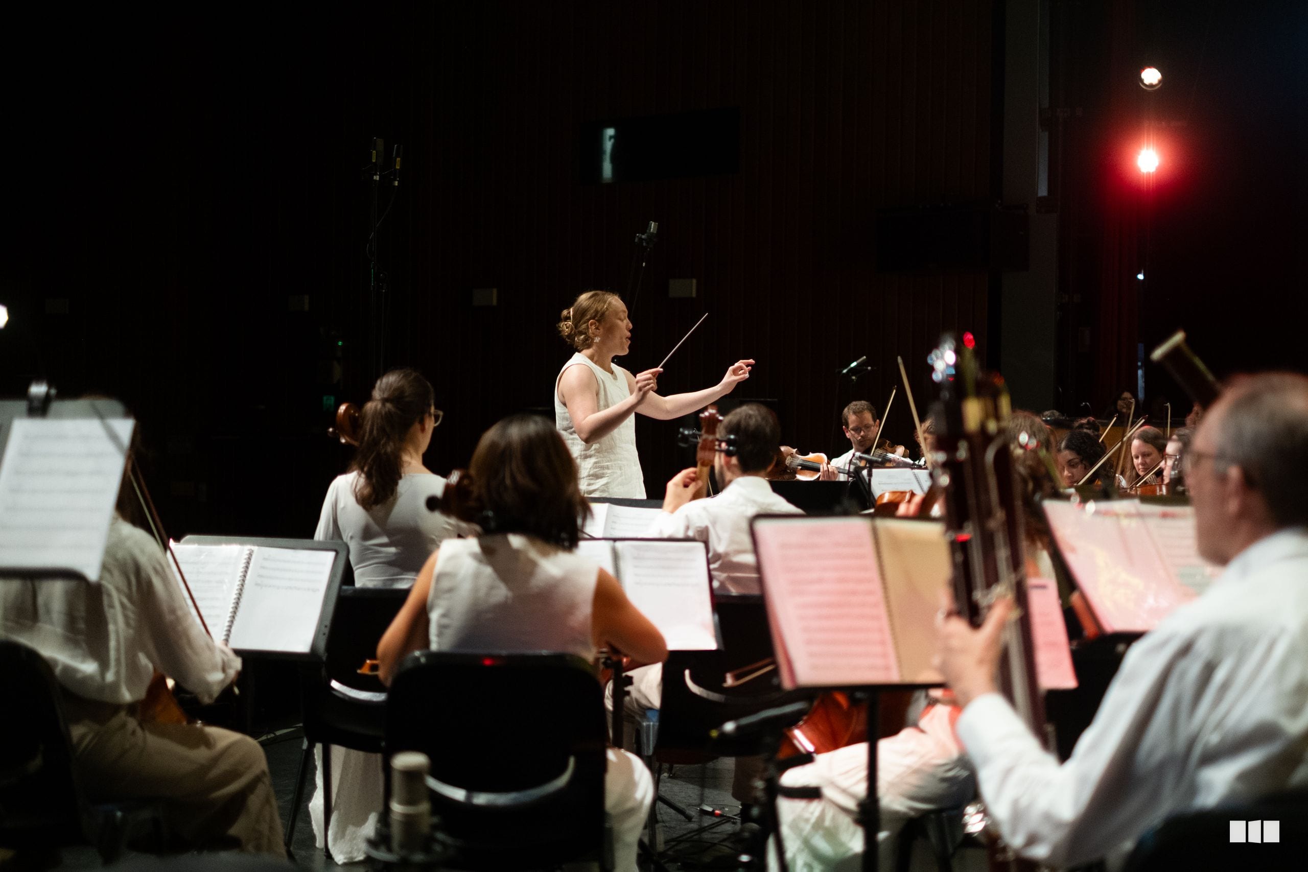 A conductor leads an orchestra during a rehearsal or performance in a concert hall with musicians reading from sheet music stands.