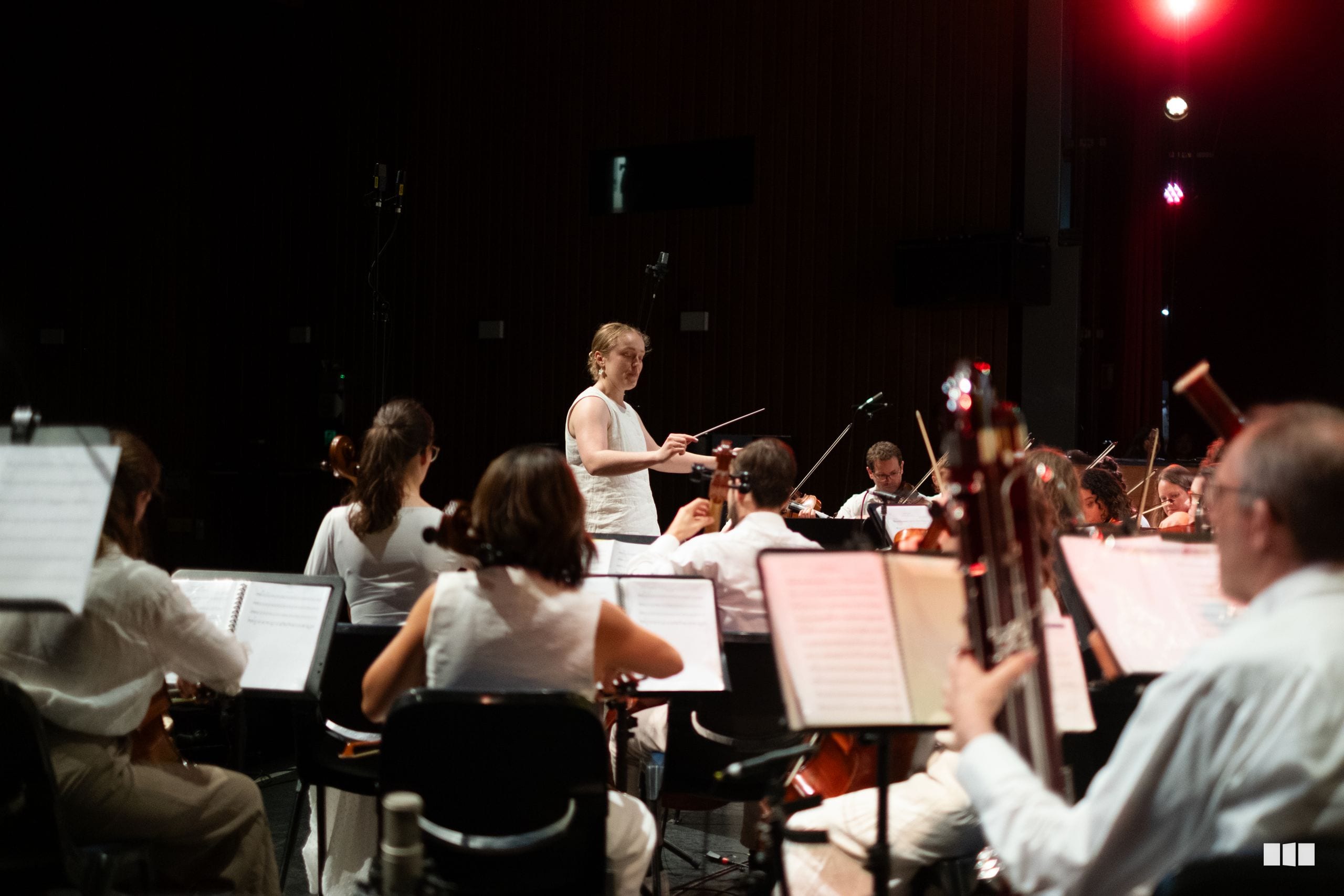A conductor leads the orchestra during a concert performance with musicians reading sheet music in the dimly lit venue.