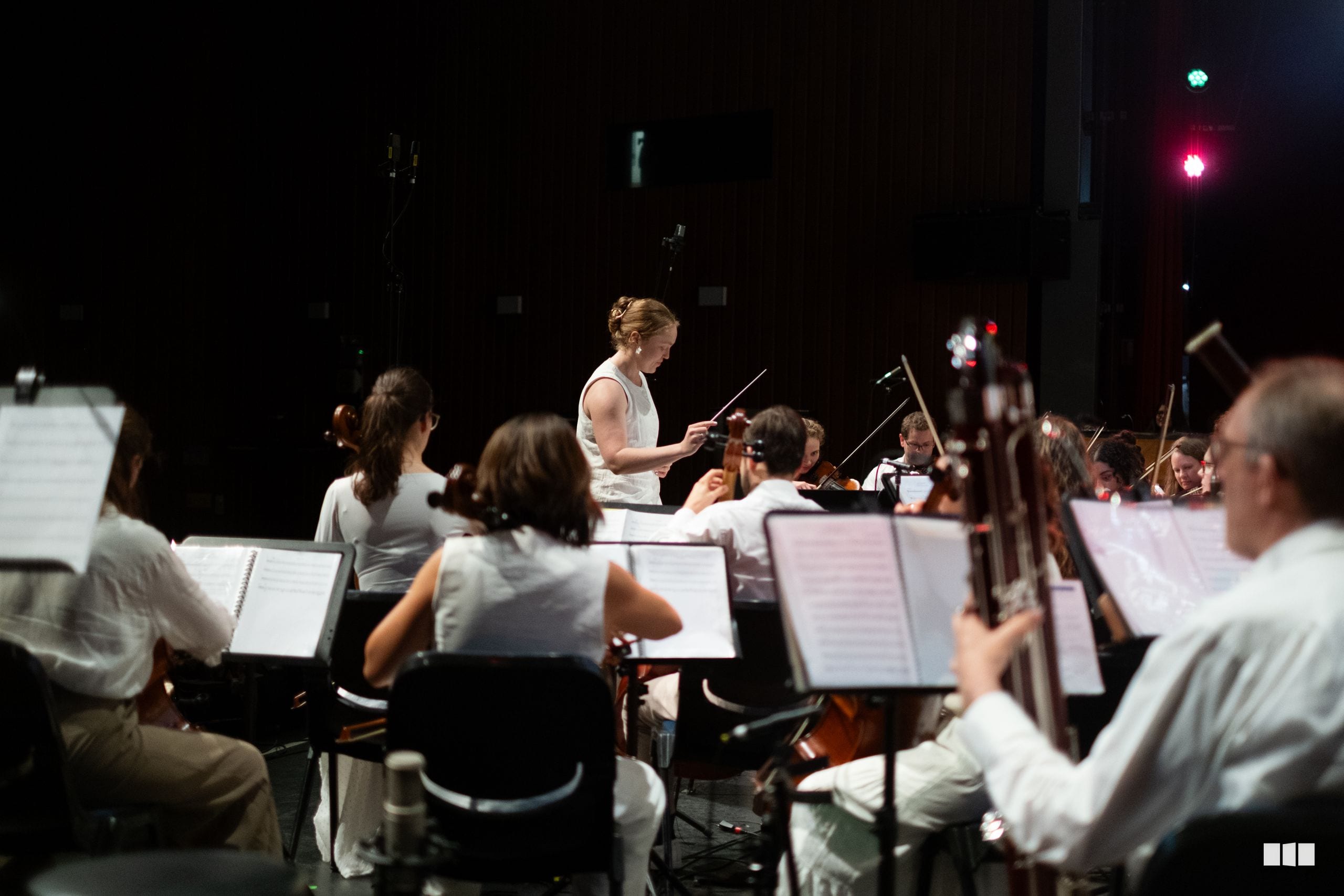 A conductor leads an orchestra during an evening performance, with musicians holding sheet music and playing various instruments under stage lighting.