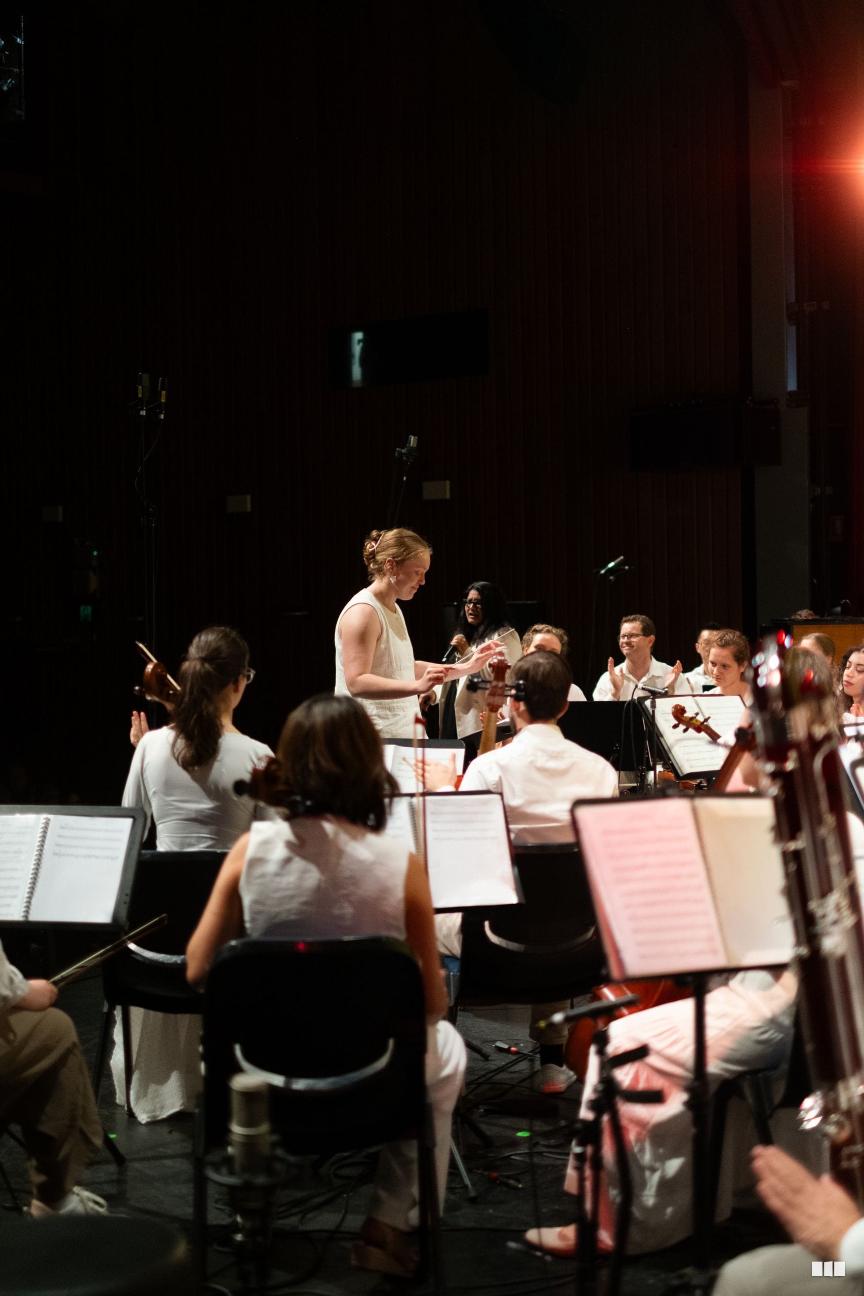 A conductor leads the orchestra during a rehearsal or performance, with musicians seated with sheet music in the darkened concert hall.