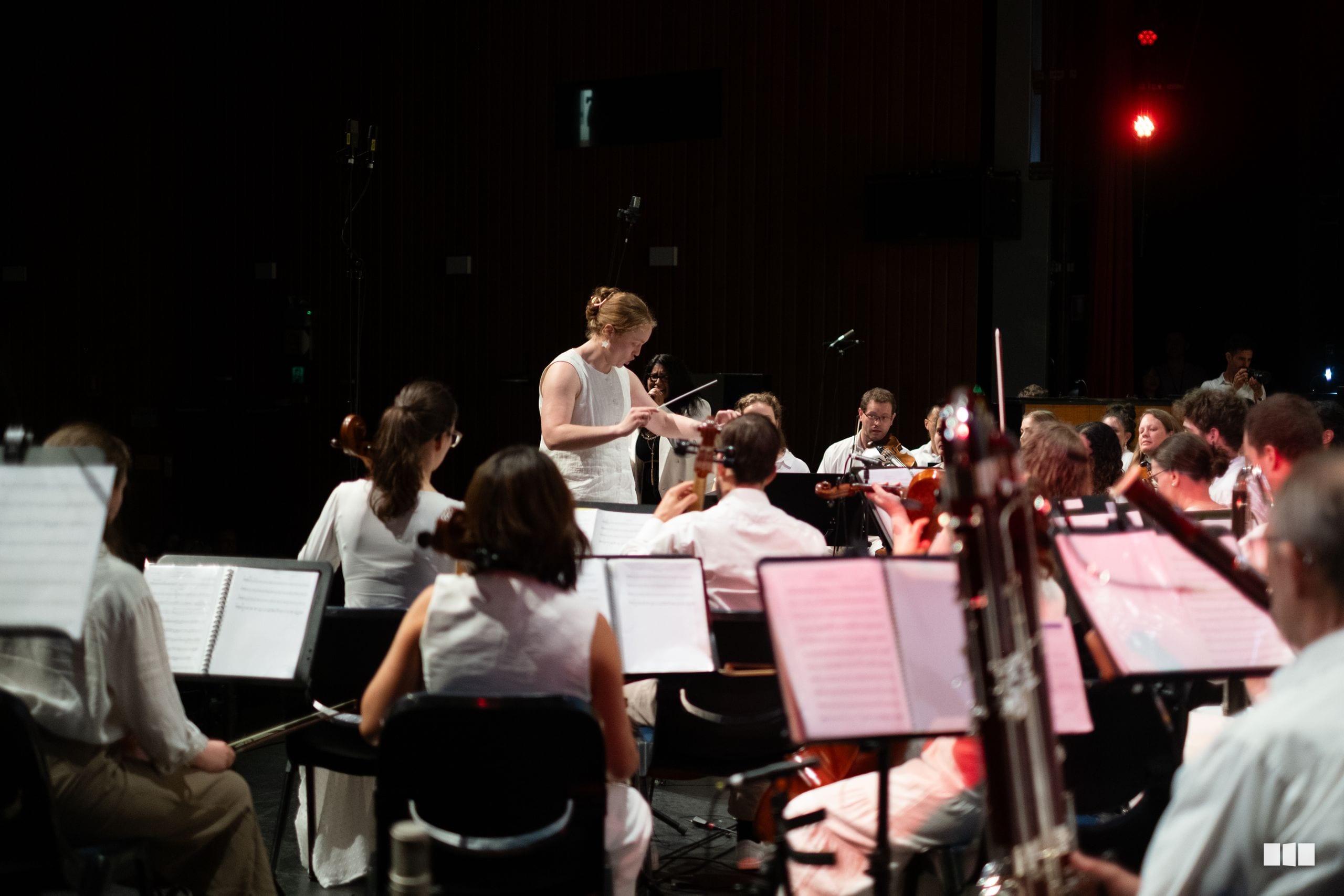 A conductor leads an orchestra during a rehearsal or performance in a darkened venue with music stands illuminated.