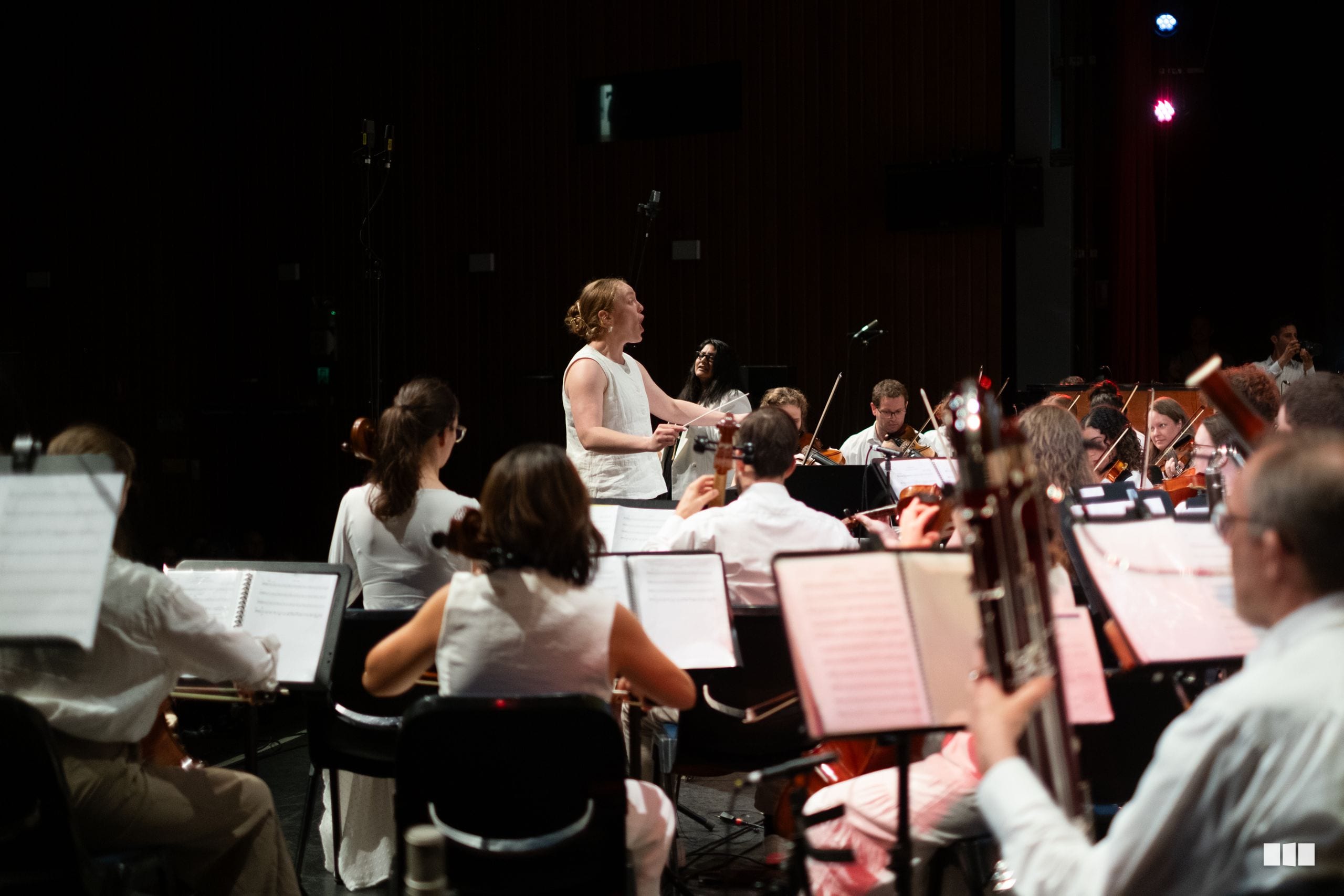 A conductor leads a string section during an orchestral rehearsal or performance on stage.