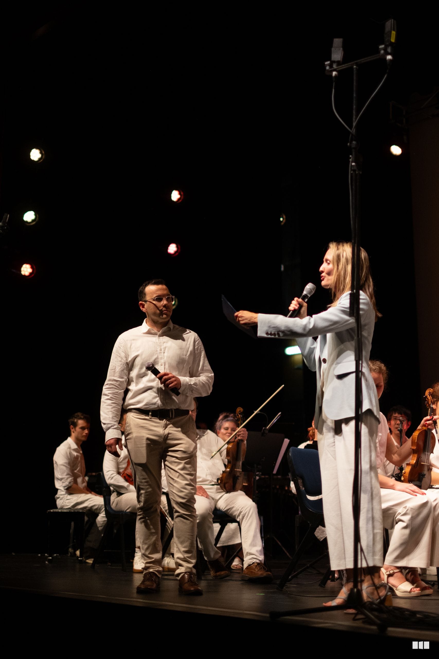A woman in a white blazer speaks at a microphone while a man in beige clothing listens during an orchestra performance on stage.