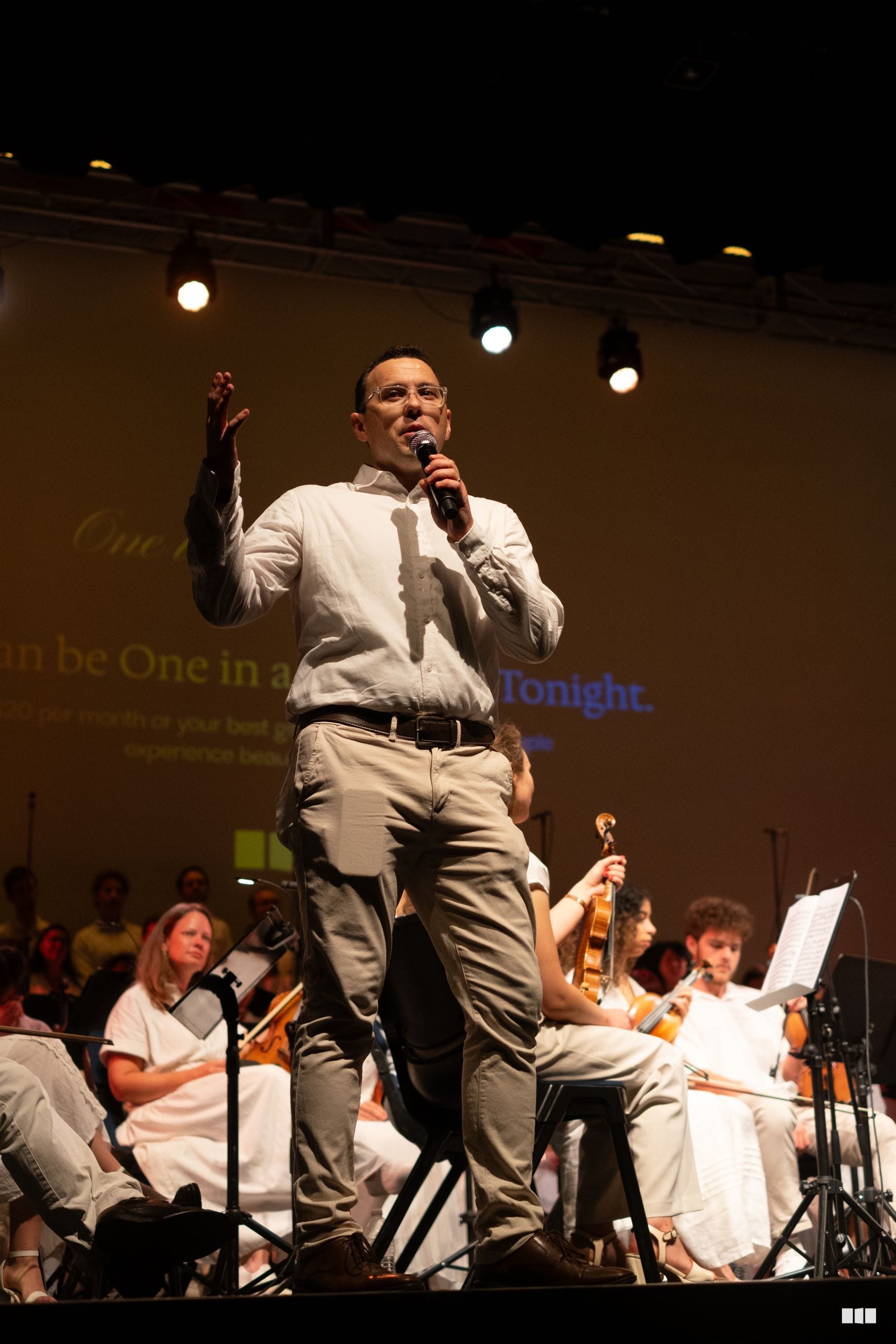 A male soloist speaks passionately to the audience while accompanied by string musicians during a performance.