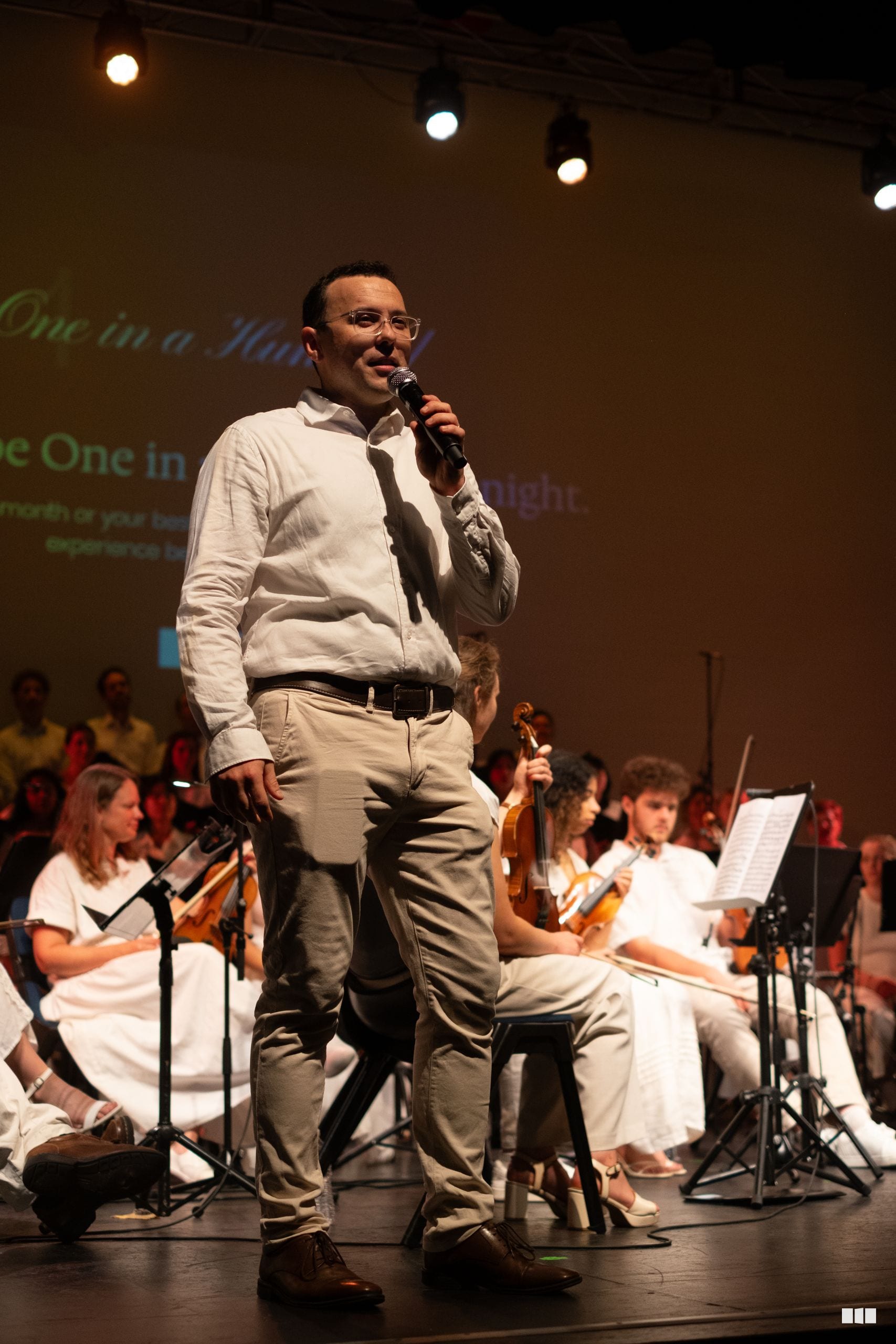 A speaker addresses the audience while seated orchestra members accompany on stage during a performance.