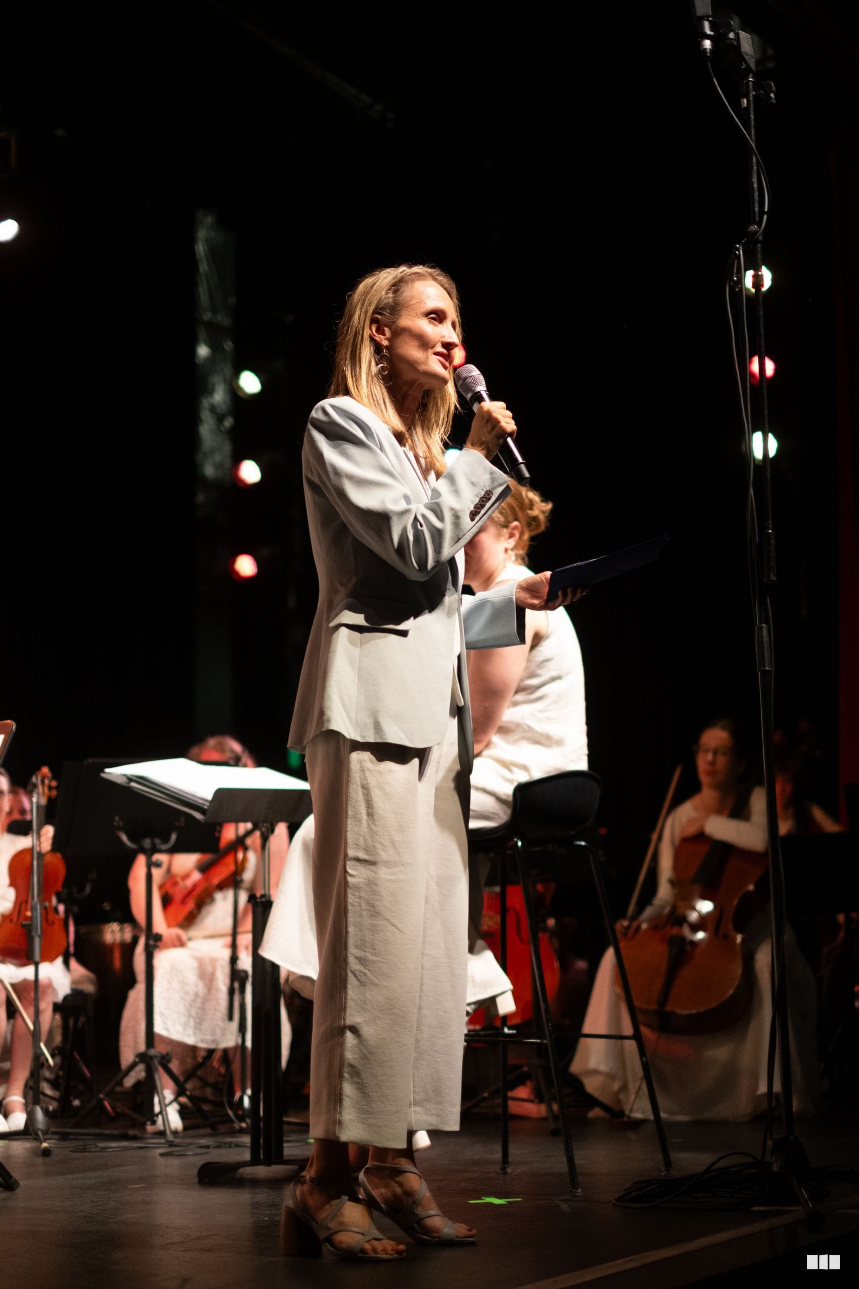 A woman in a cream linen suit speaks at a microphone on stage with orchestra musicians and string players visible in the background.