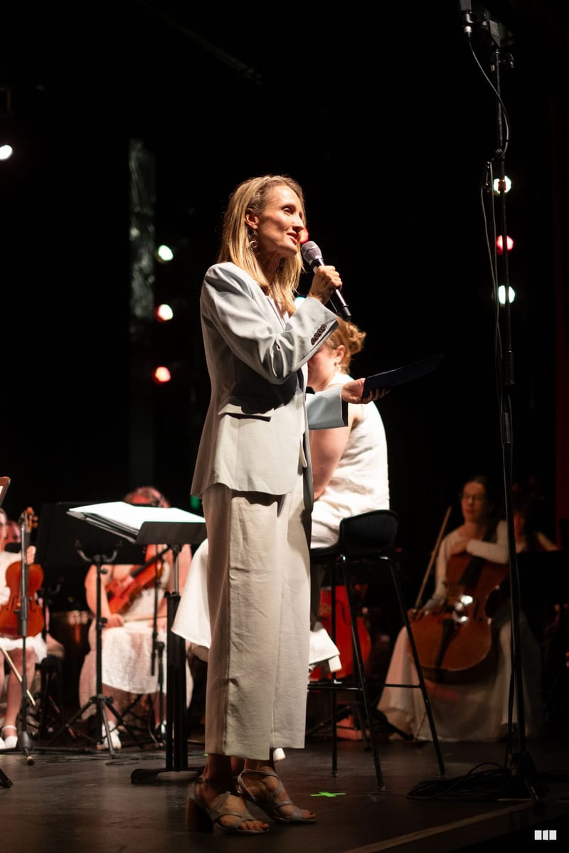 A woman in a cream linen suit speaks at a microphone on stage with orchestra musicians and string players visible in the background.