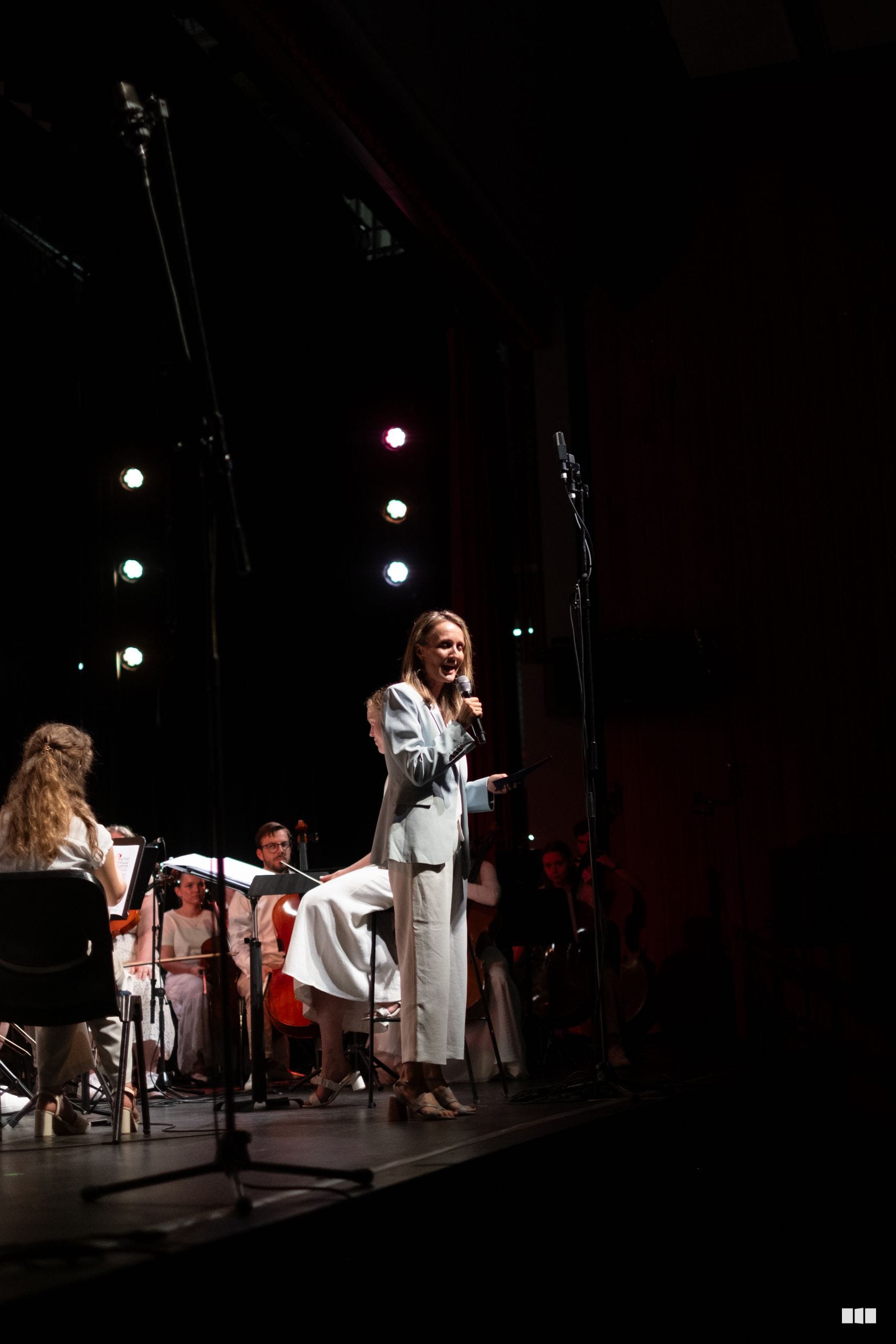 A soloist in a light blue dress performs at the microphone with orchestra members seated beside her on stage under dramatic theatrical lighting.