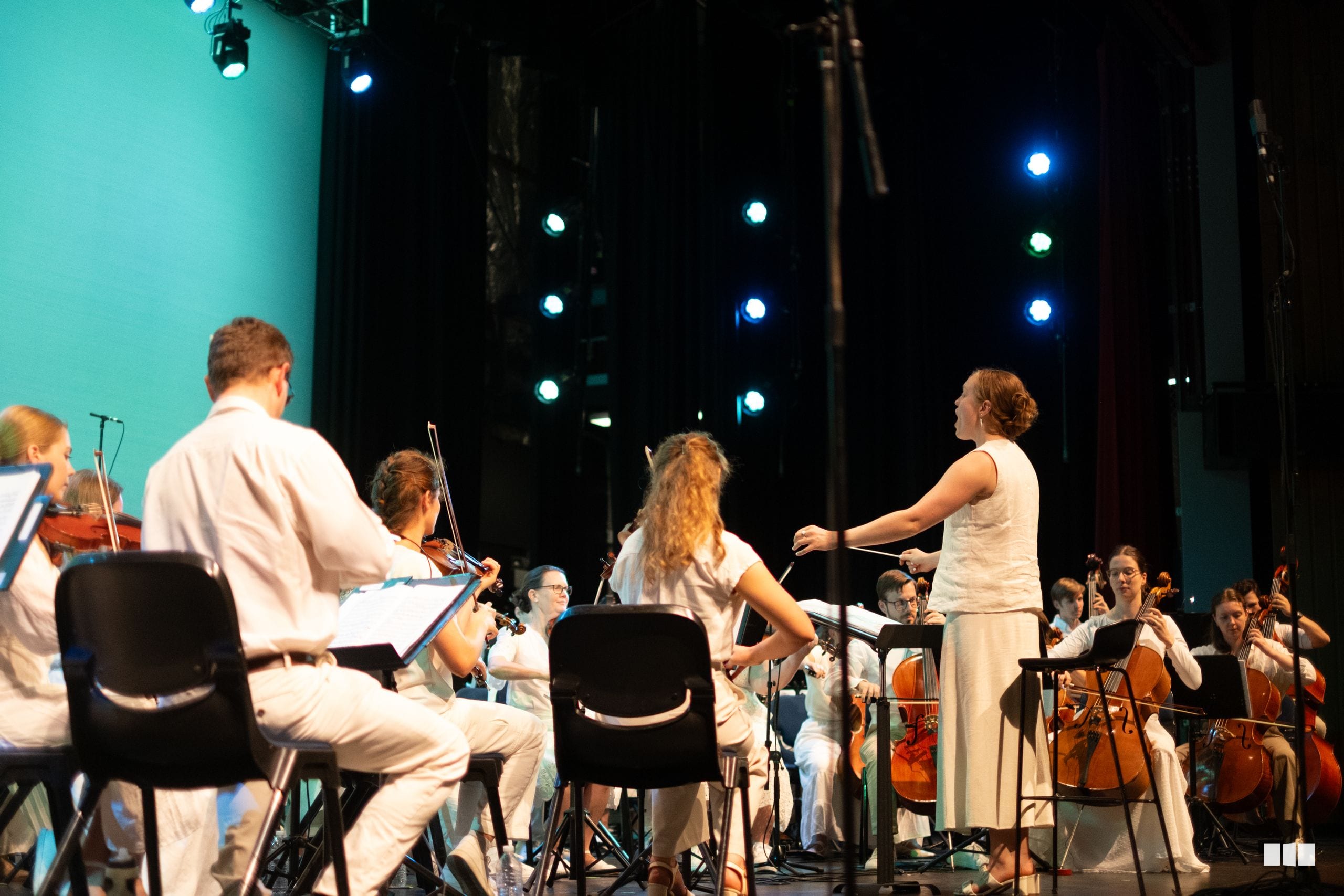 A conductor leads an orchestra with string and woodwind players during a performance on stage with blue stage lighting.