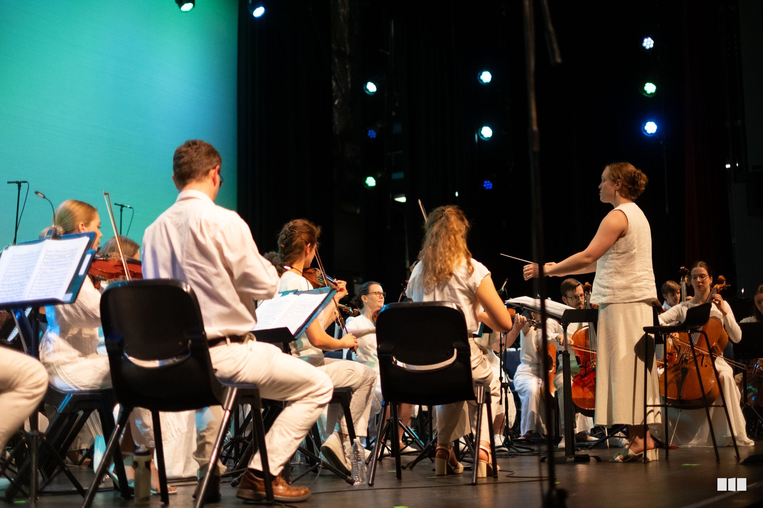 A conductor leads a youth orchestra ensemble during a performance on stage with turquoise lighting and musical scores visible.
