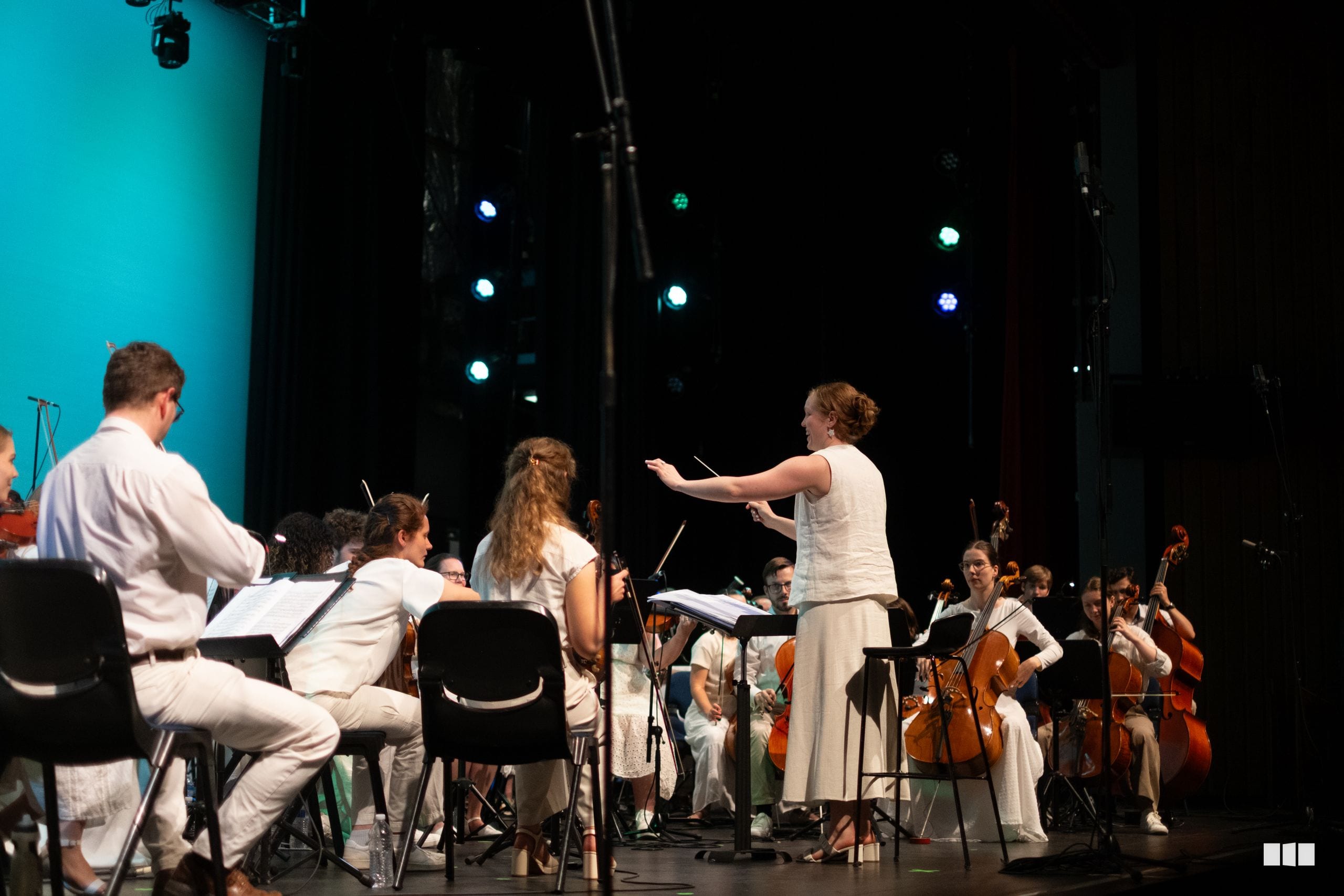 A conductor leads a string orchestra during a performance on stage with professional lighting.