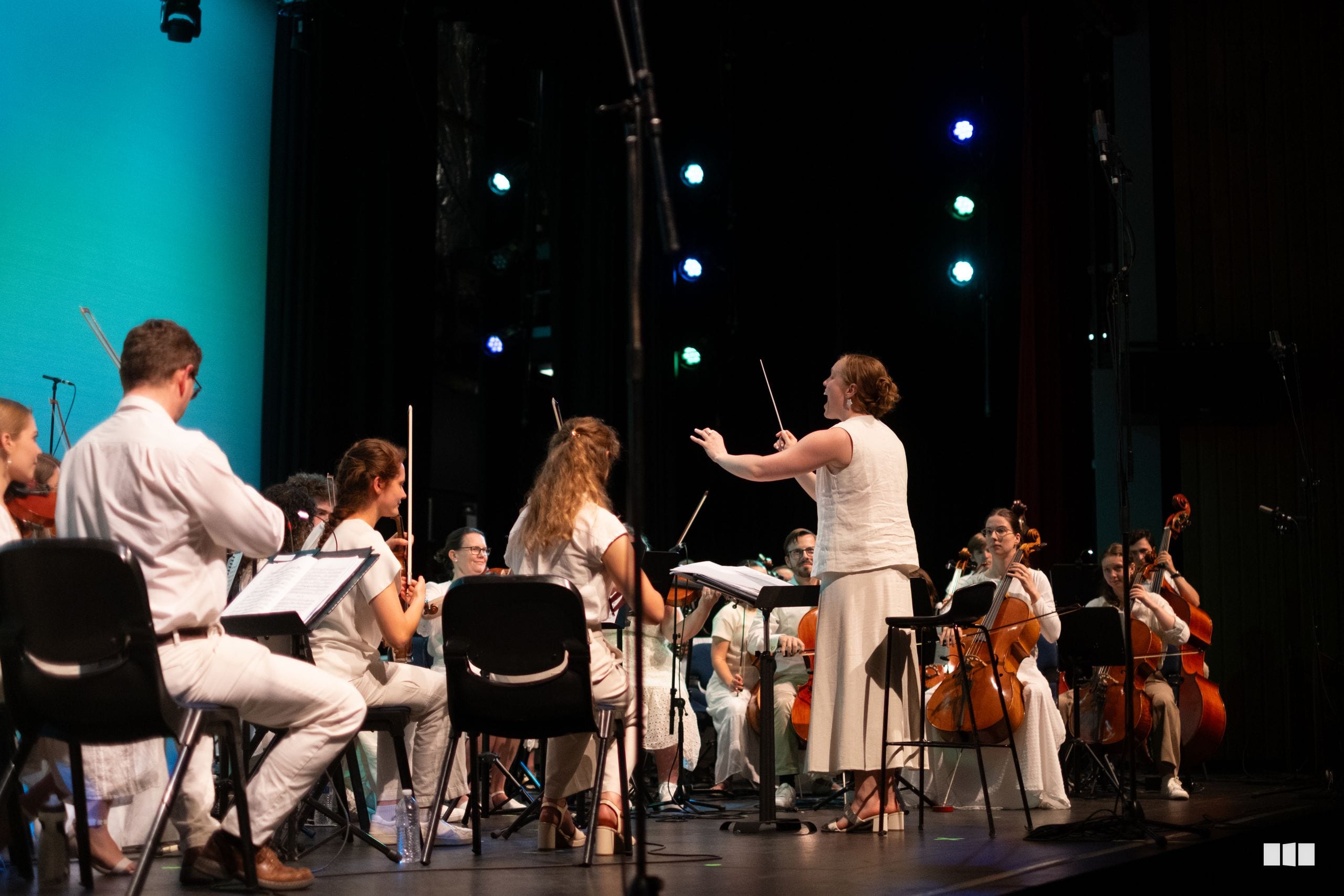 A conductor leads a youth orchestra during a performance on stage with professional lighting.