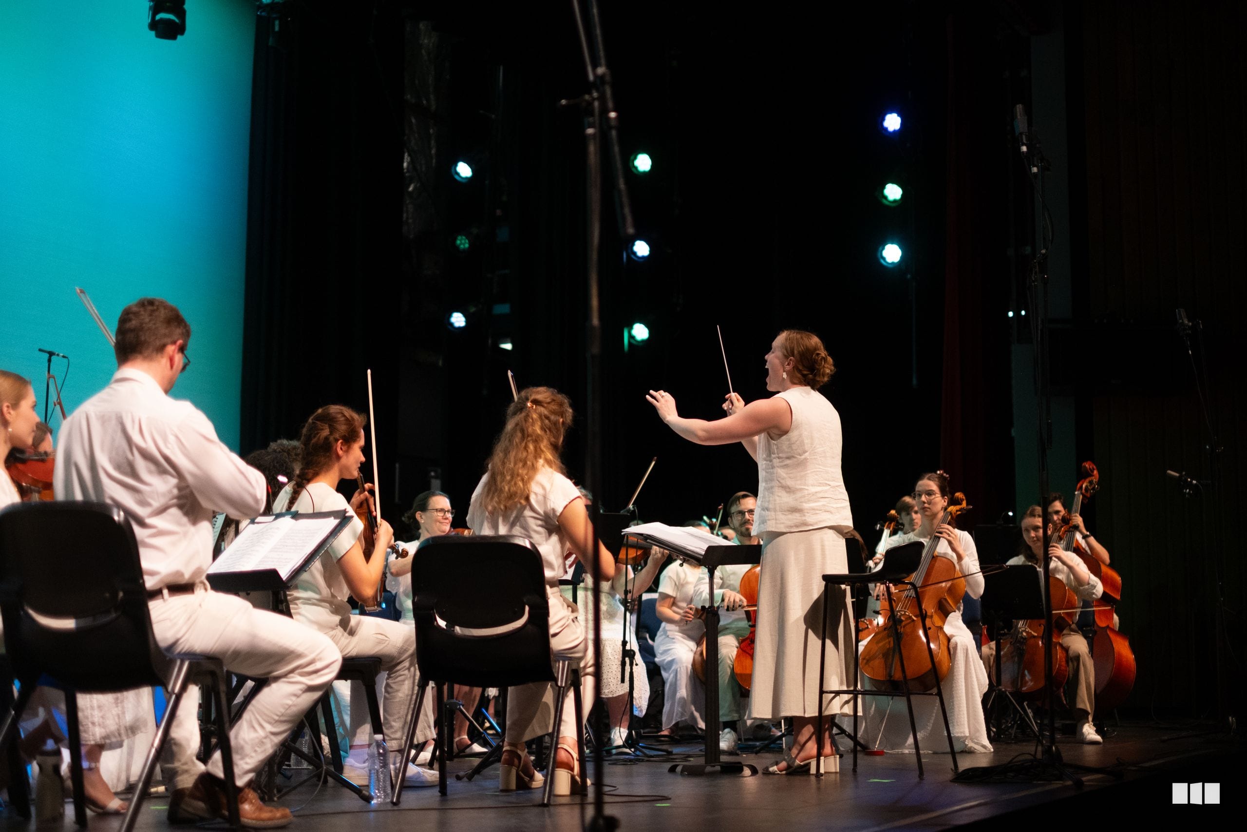 A conductor leads a string orchestra during a performance on stage with professional lighting.