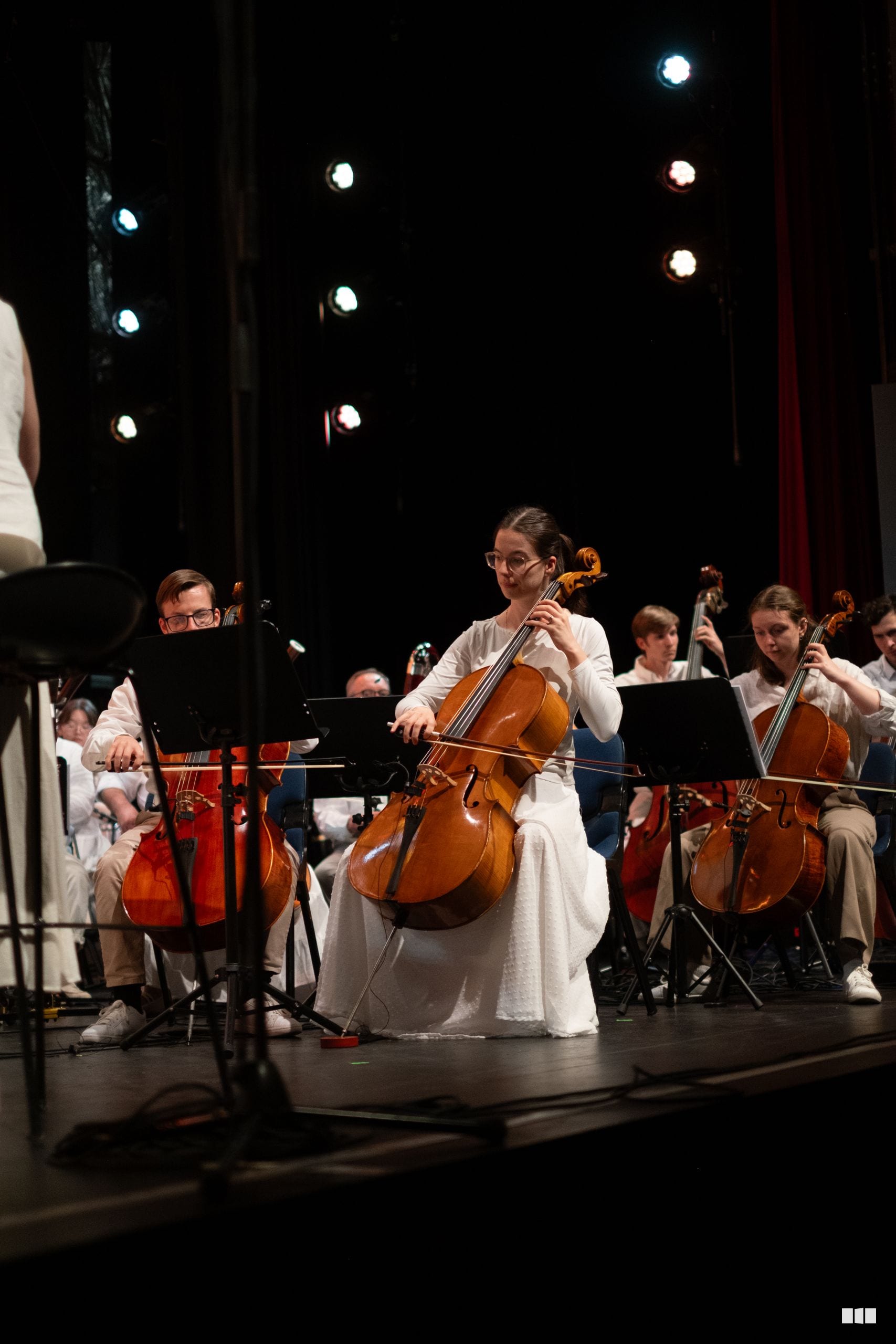 A young cellist in white performs as a soloist with the orchestra during a concert under stage lighting.