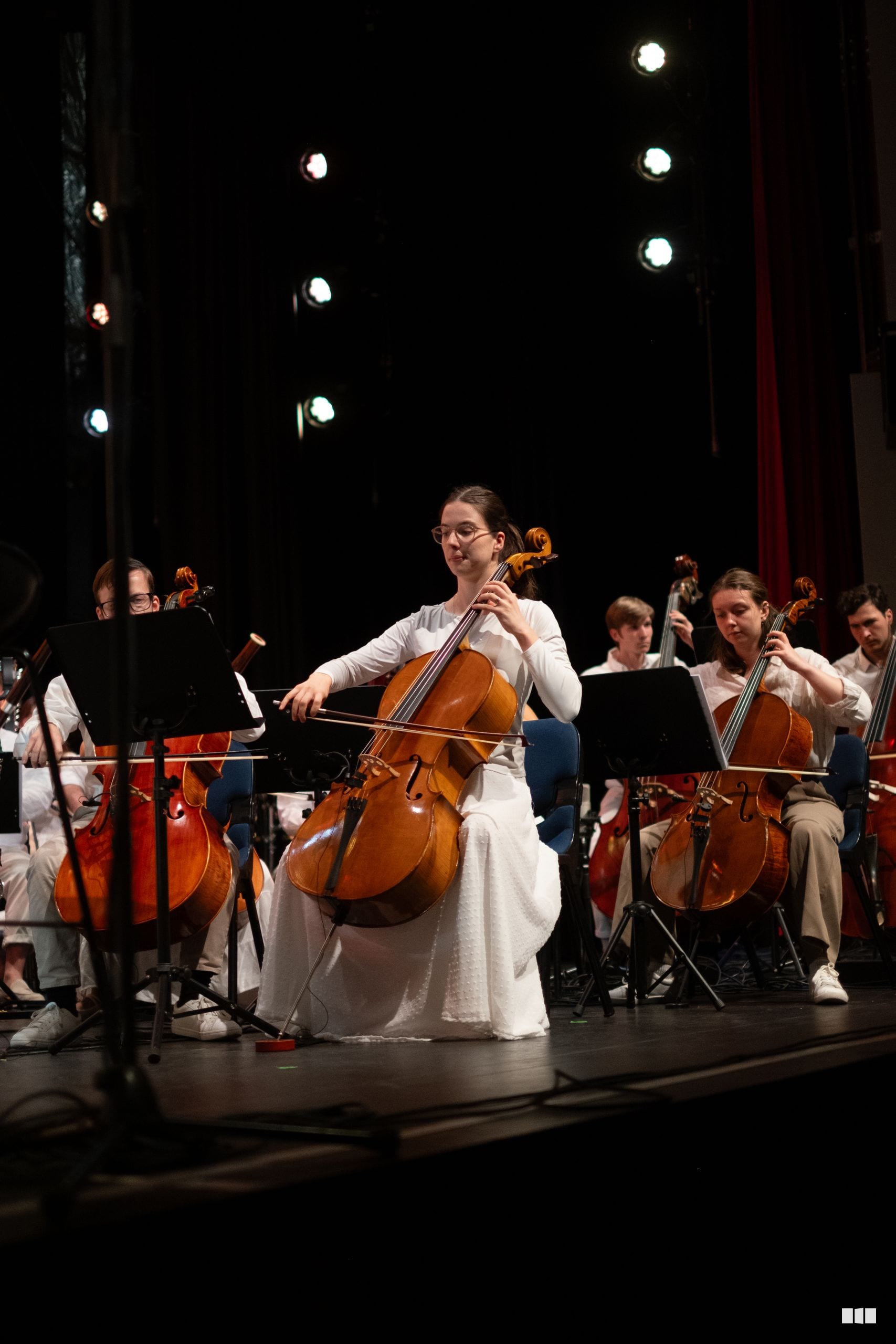 A cellist in white performs as a soloist with the string section during an orchestral concert under stage lighting.