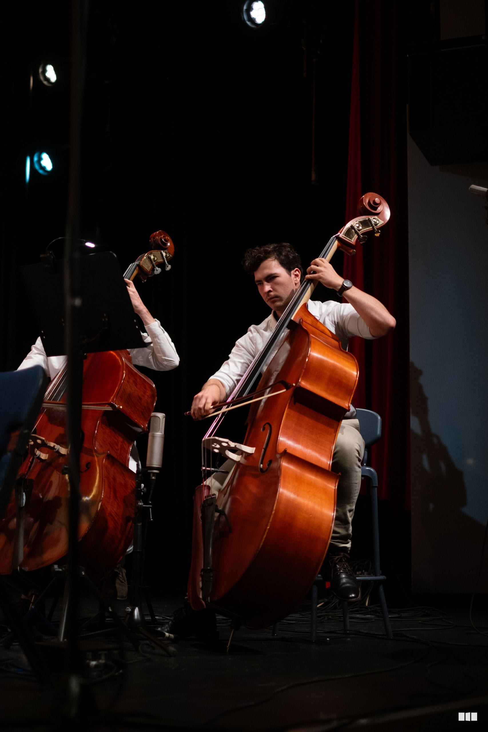 Two cellists perform together on stage with professional stage lighting and red curtain backdrop.