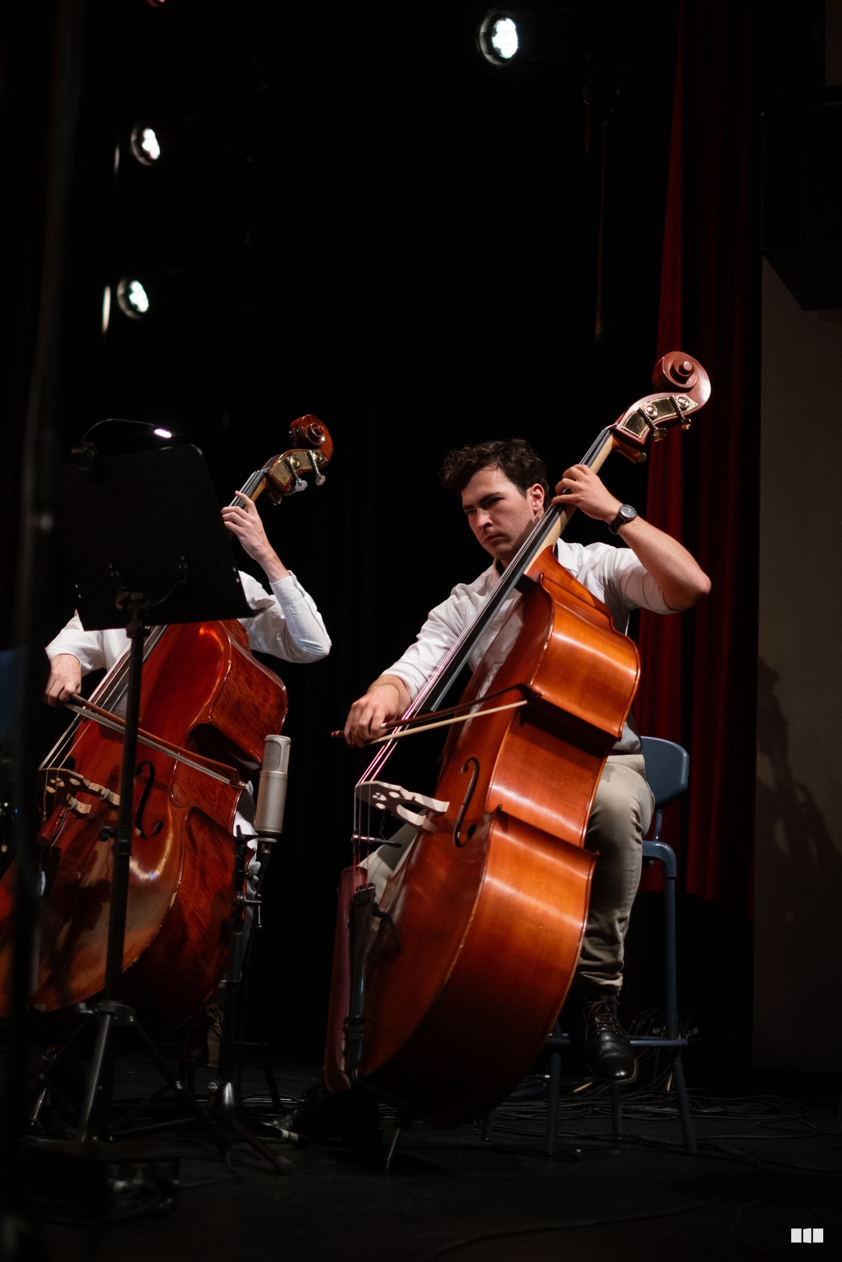 Two cellists perform together on stage, with one musician in focus playing a warm-toned wooden cello under professional stage lighting.