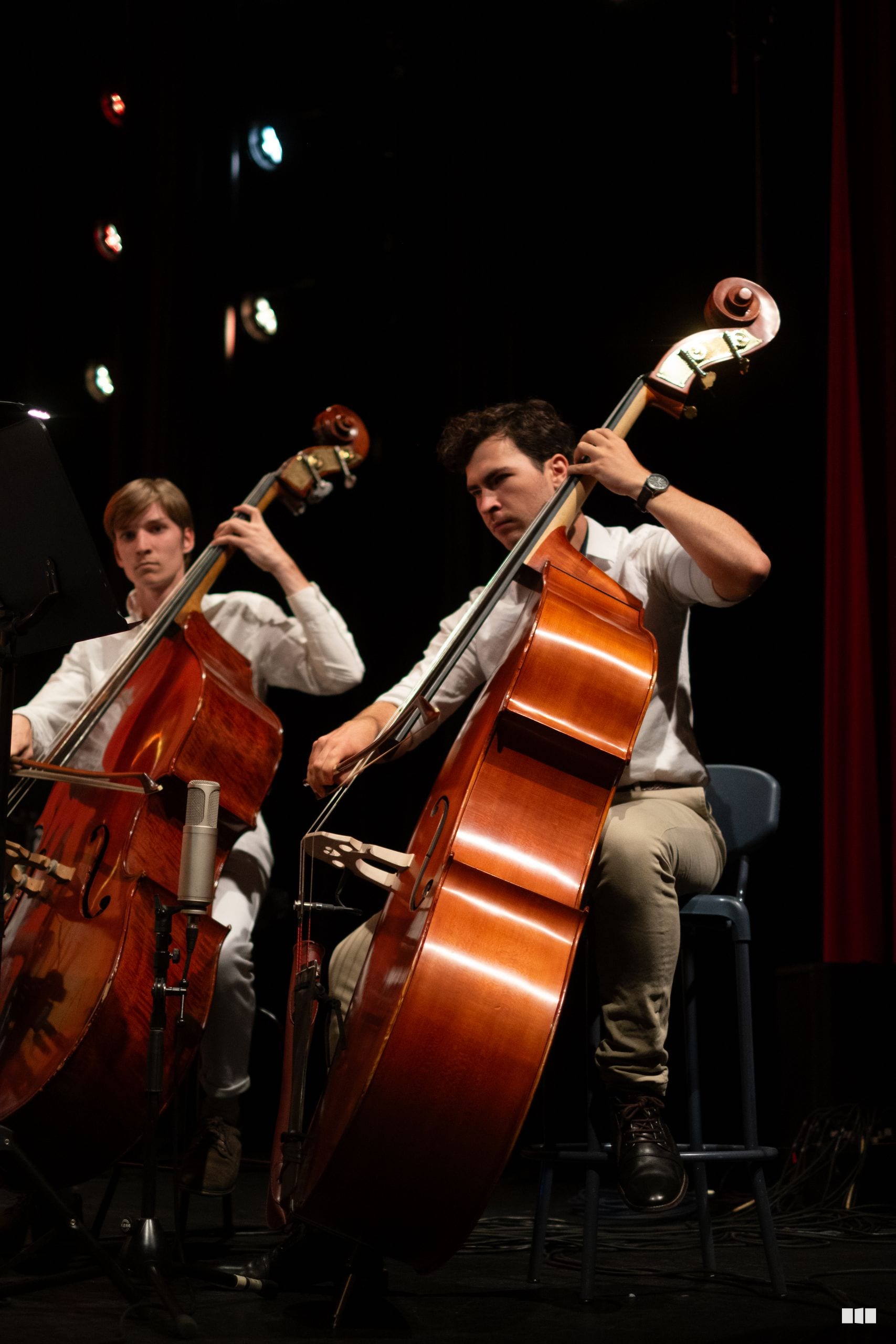 Two cellists perform together on stage with warm stage lighting and a red curtain backdrop.