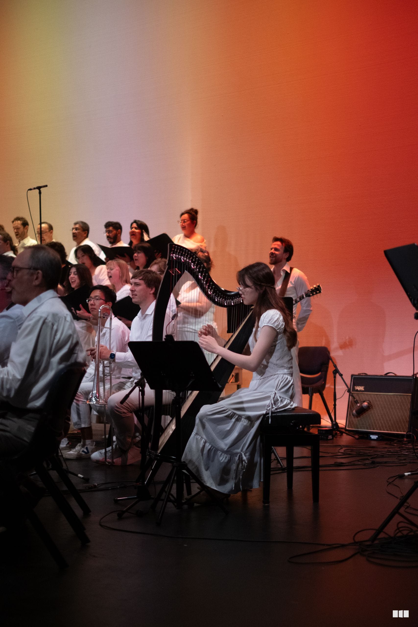 A mixed ensemble of orchestra and choir members perform together on stage under warm amber lighting, with harp, brass, and vocal soloists featured prominently.