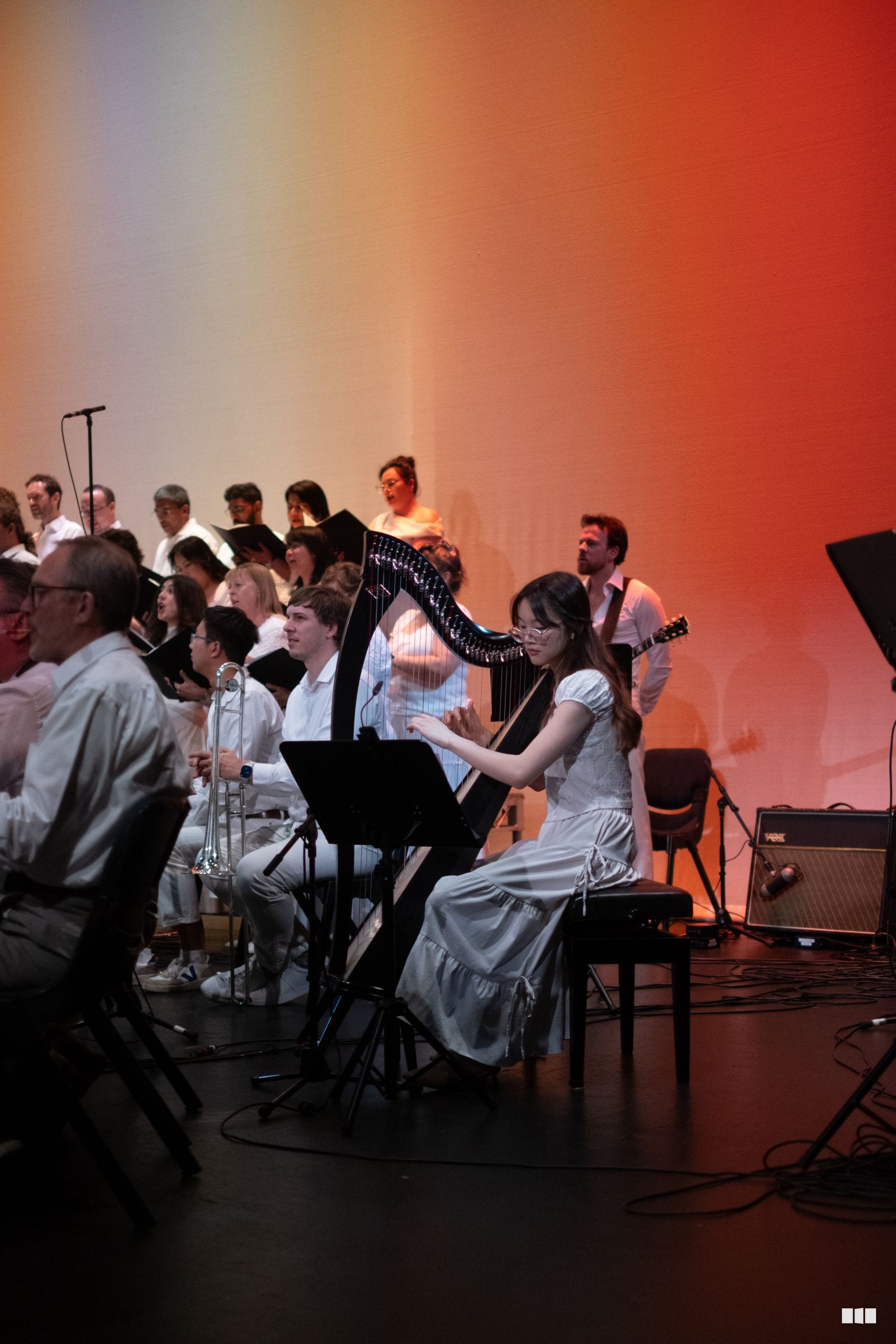 A harpist in a light blue dress performs alongside orchestra and choir members on stage under warm lighting during a concert.