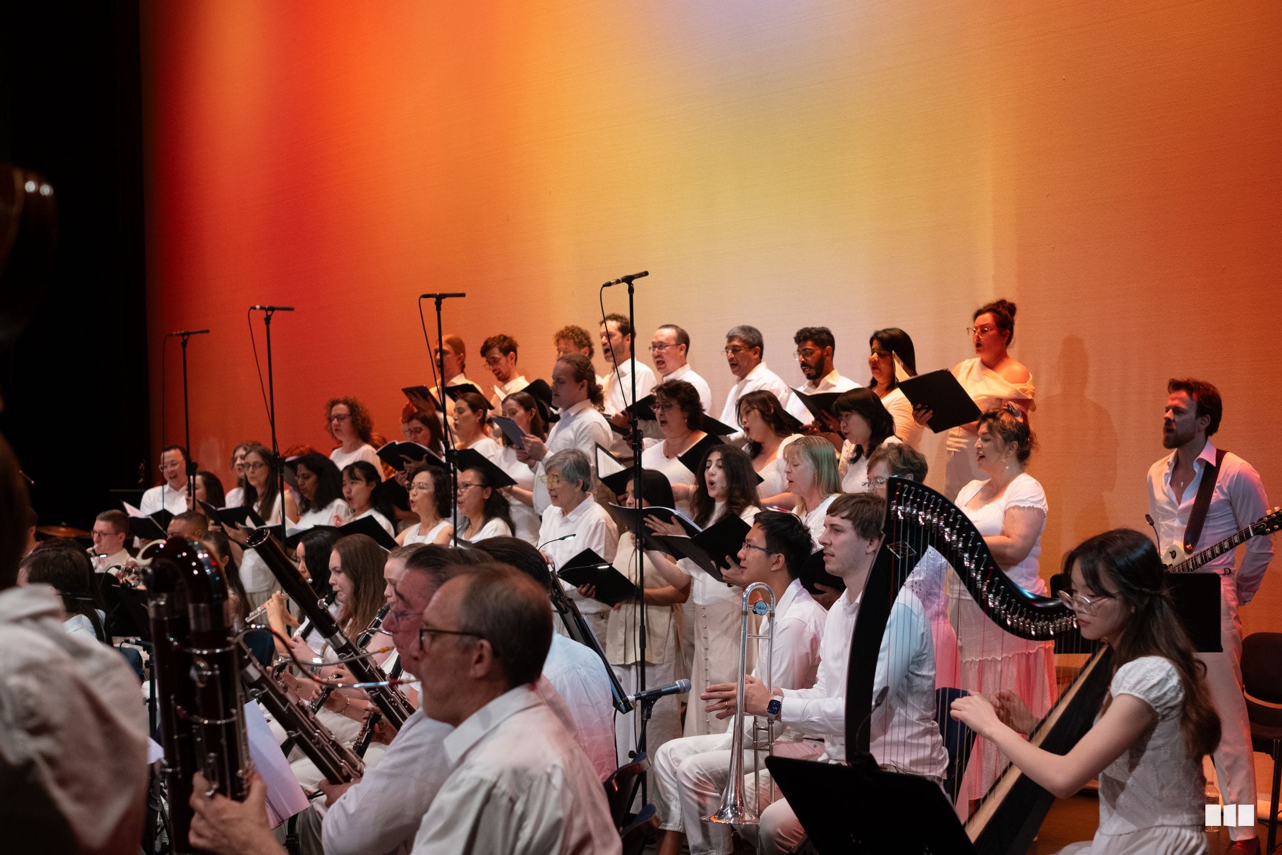 A large choir and orchestra ensemble perform together on stage with warm orange lighting during a concert.