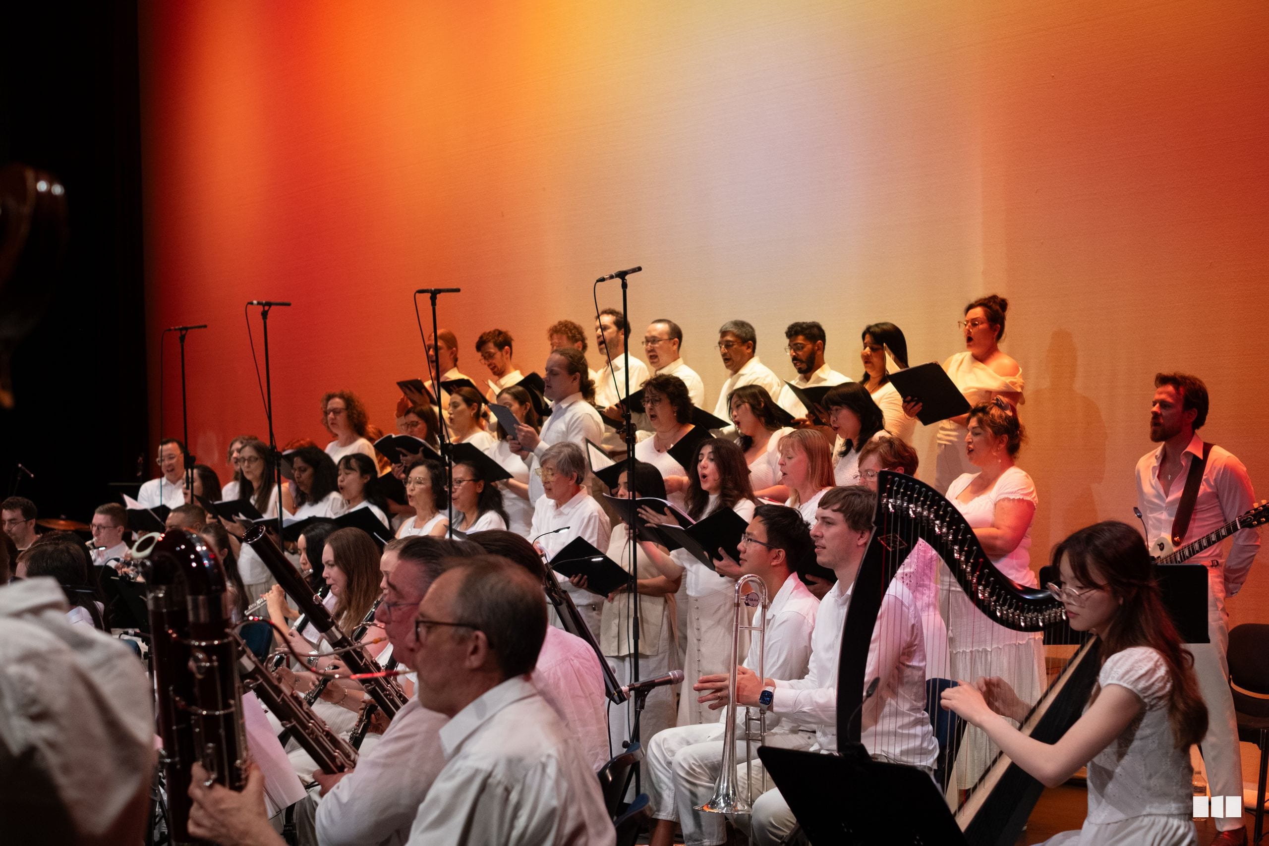 A large ensemble of orchestra and choir members perform together on stage with warm orange lighting, featuring brass, woodwind, and string players alongside singers holding sheet music.