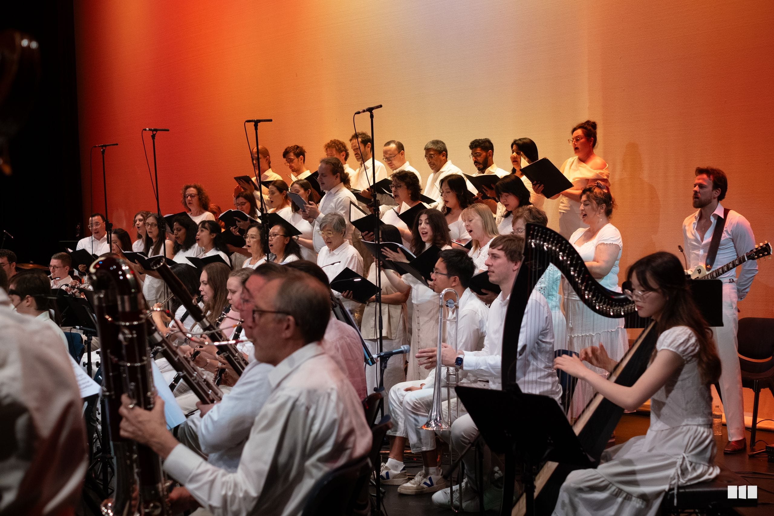 A mixed choir and orchestra perform together on stage with warm orange stage lighting, featuring singers in white shirts and instrumentalists including brass and harp players.