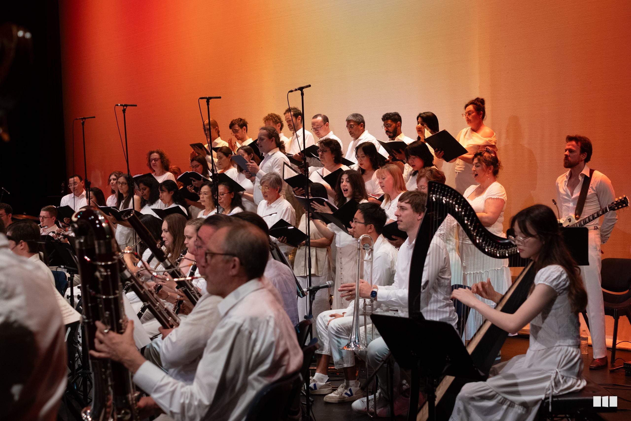 A combined orchestra and choir performs together on stage under warm orange lighting, with musicians holding instruments and singers reading from scores.