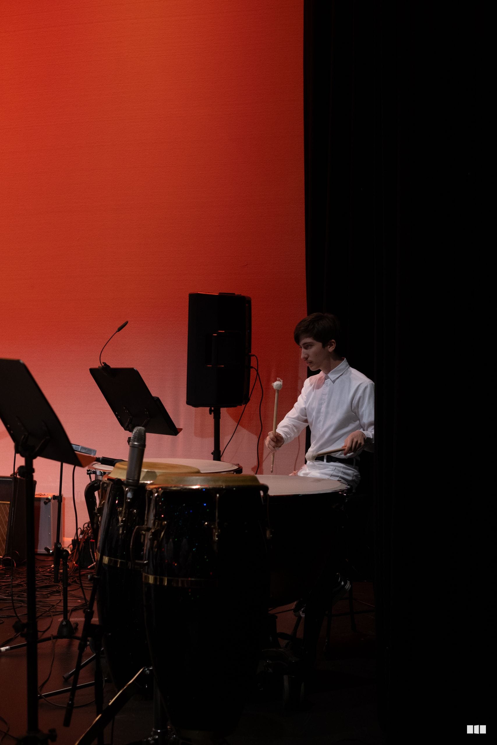 A young percussionist performs at the drums during an orchestral concert under vibrant red stage lighting.