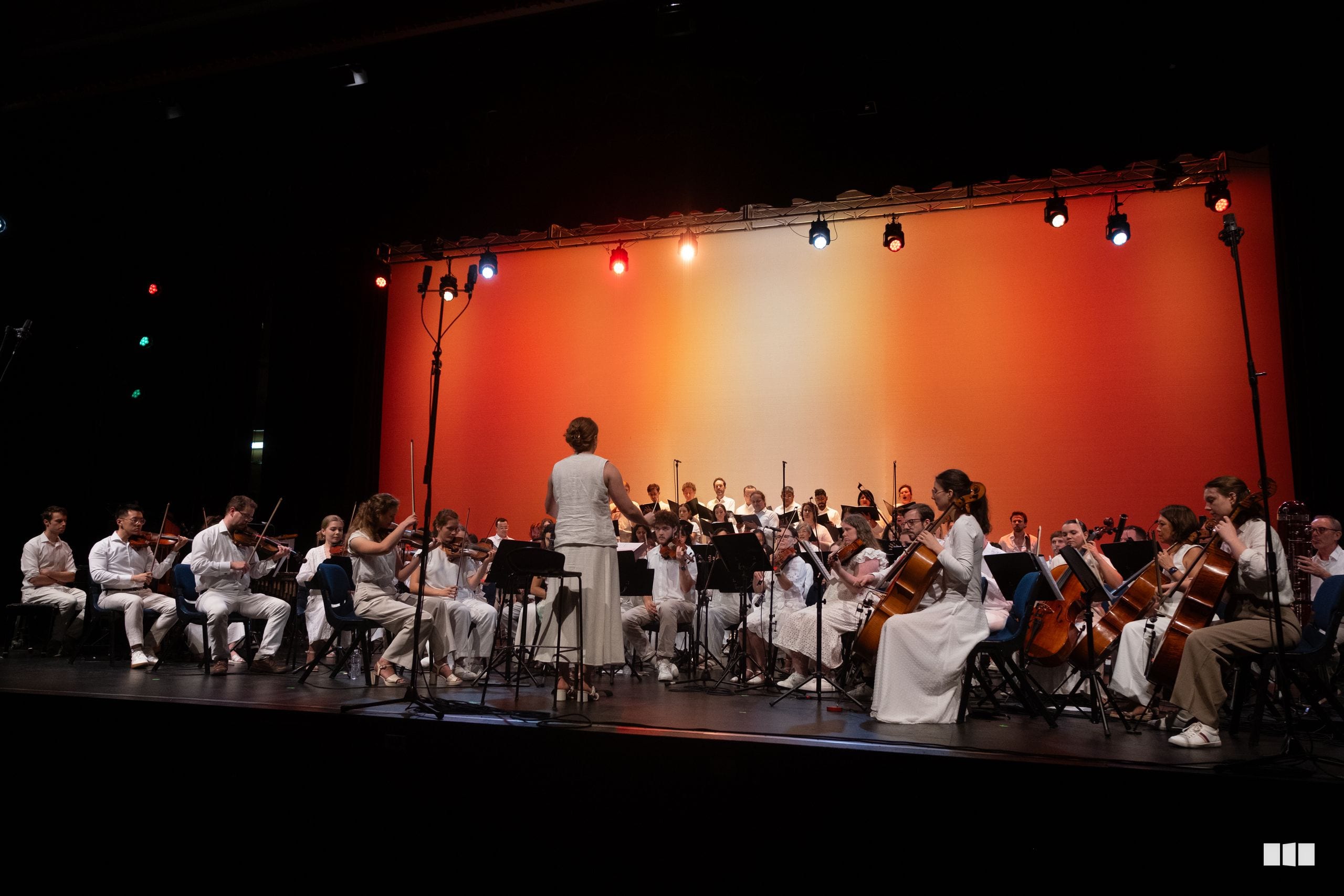 A conductor leads a large orchestra performing on stage with an orange-red backdrop and professional stage lighting.