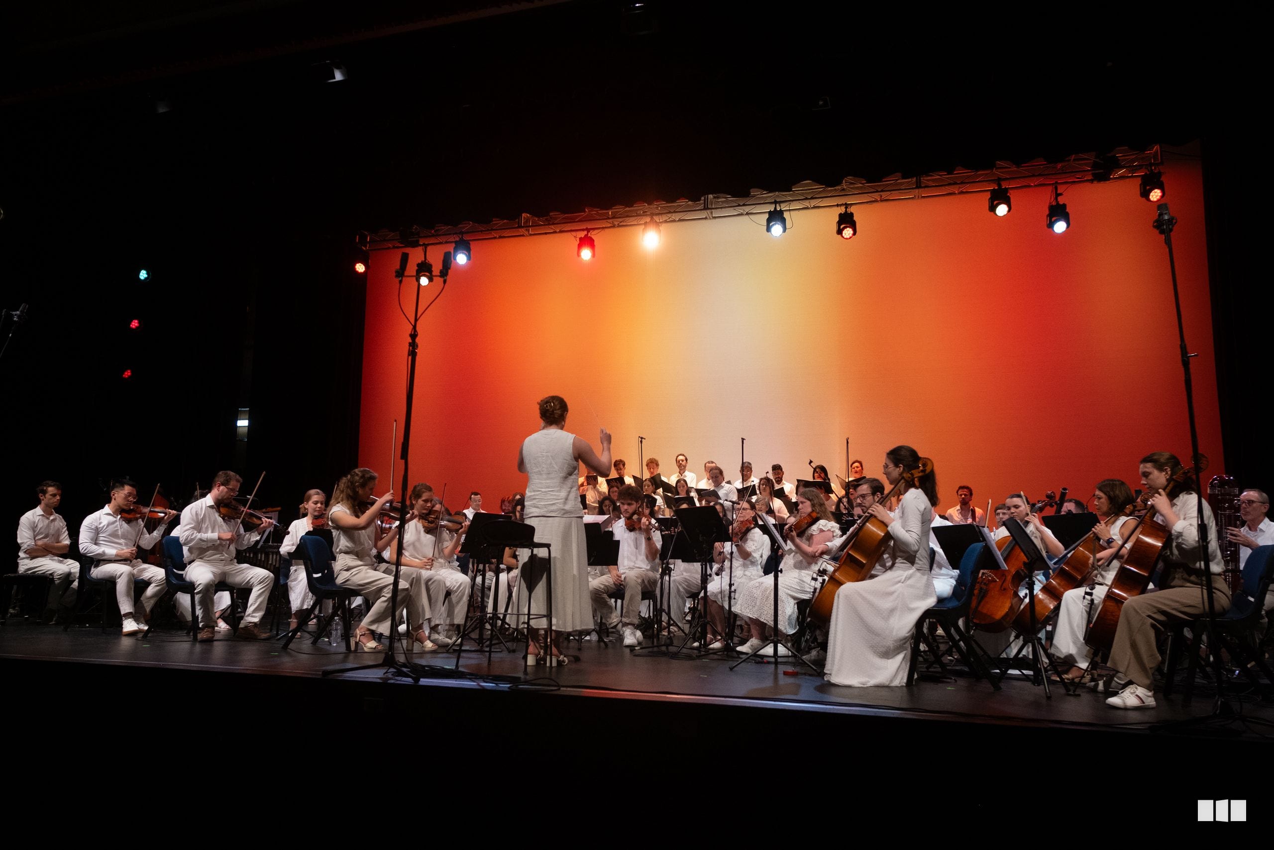 A conductor leads a large orchestra dressed in white against a vibrant orange-lit backdrop during a concert performance.