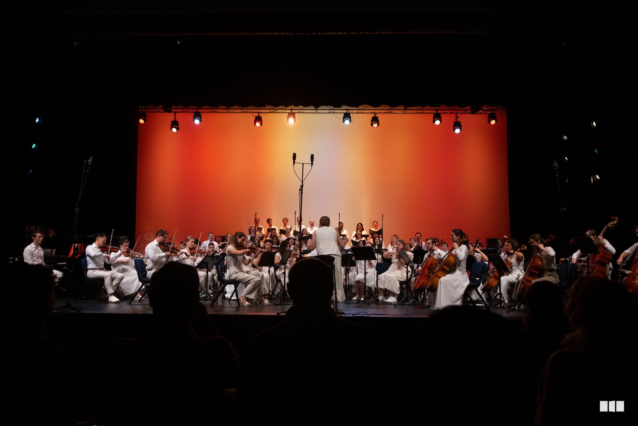 A conductor leads a full orchestra in white attire performing on stage with an orange backdrop and theatrical lighting.
