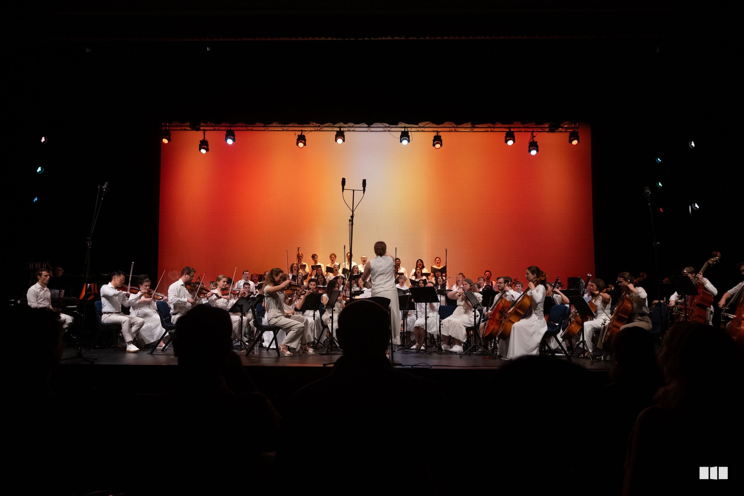 A conductor leads a full orchestra with string, woodwind, and brass sections performing on stage under warm orange lighting.