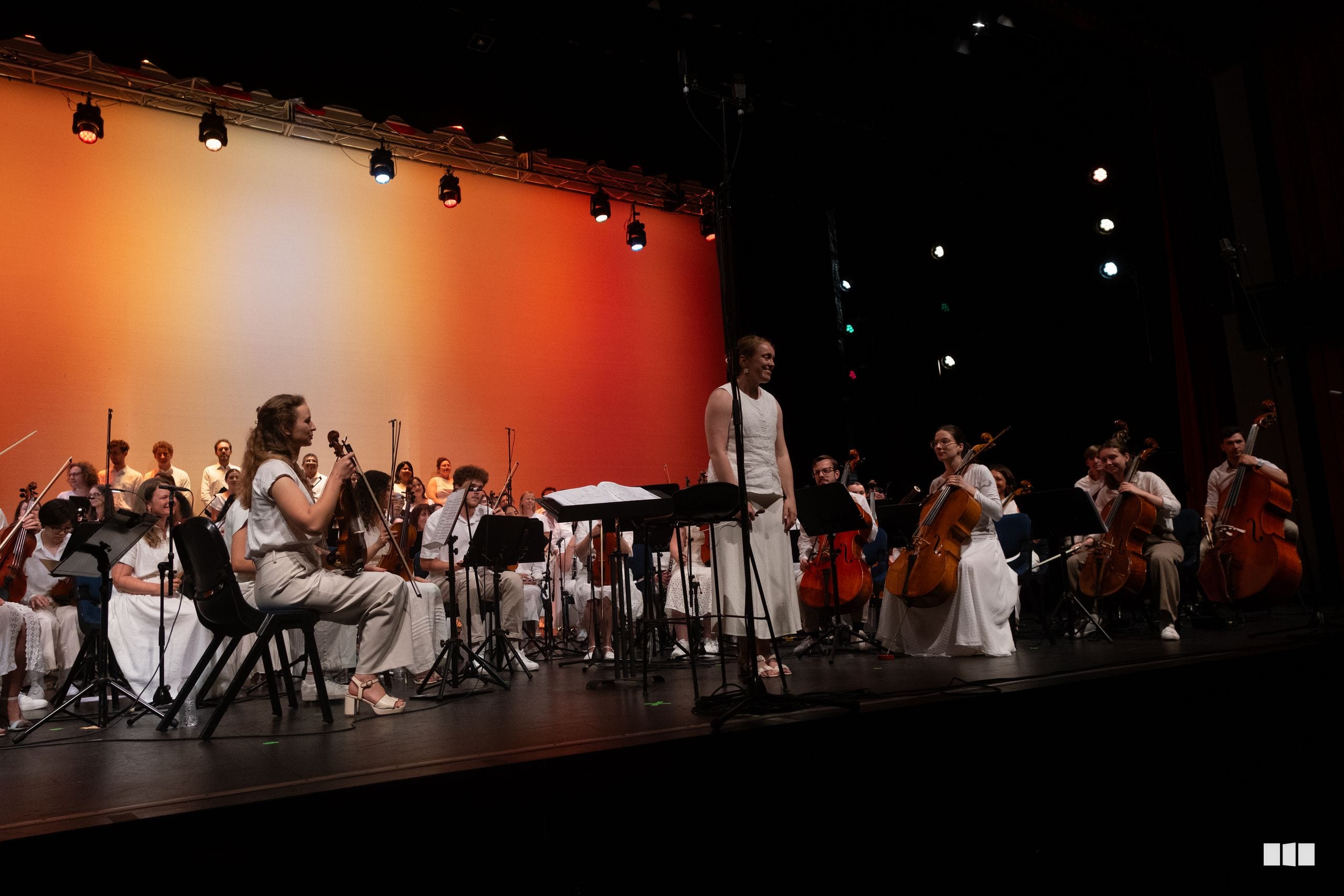 A conductor leads a chamber orchestra performance on stage with string and wind players dressed in white against a vibrant orange-lit backdrop.