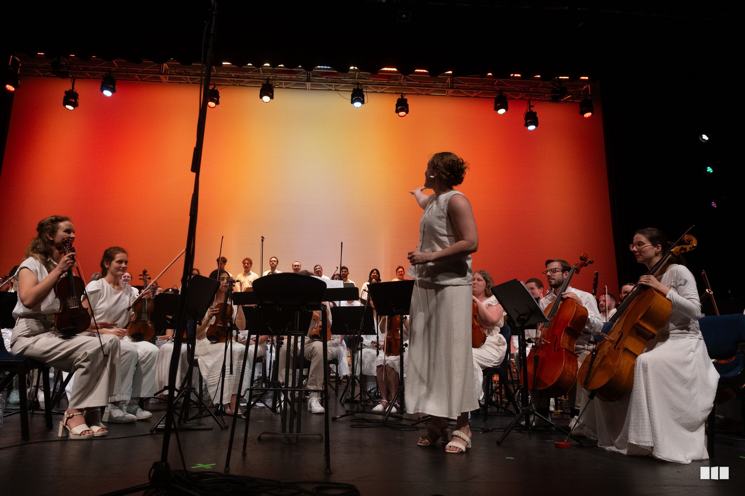 A conductor leads an orchestra of musicians dressed in white during a performance against a vibrant orange backdrop.