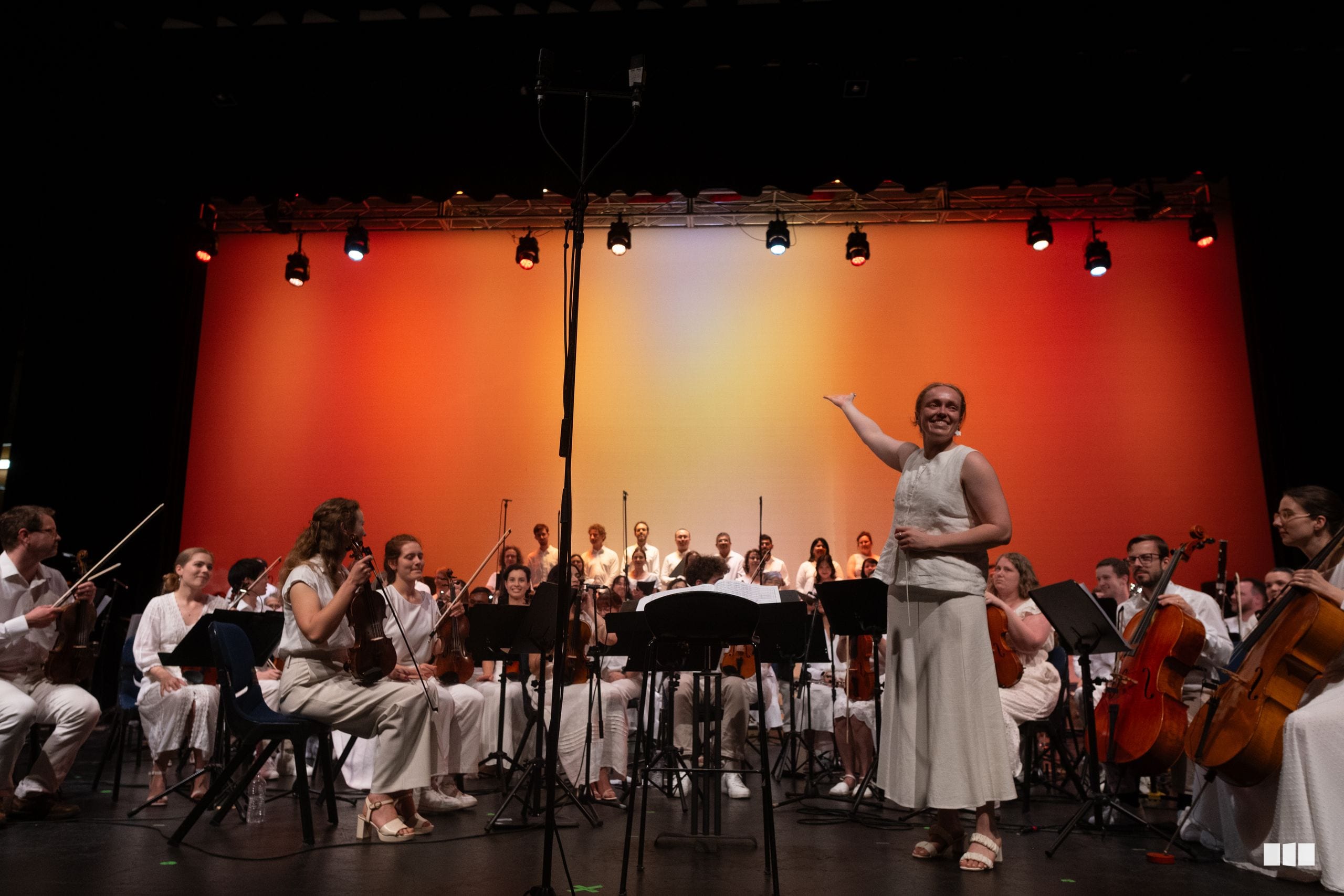 A conductor leads a full orchestra in performance with musicians dressed in light-colored attire against a vibrant orange-red illuminated backdrop.