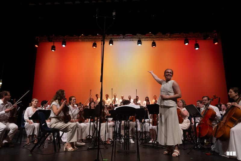 A conductor leads a full orchestra in performance with musicians dressed in light-colored attire against a vibrant orange-red illuminated backdrop.