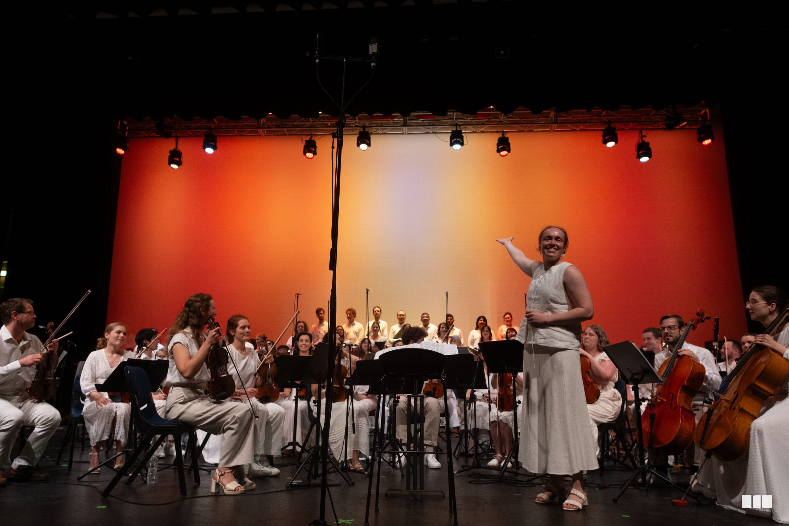 A conductor leads a large orchestra during a performance on stage with warm orange and yellow lighting.