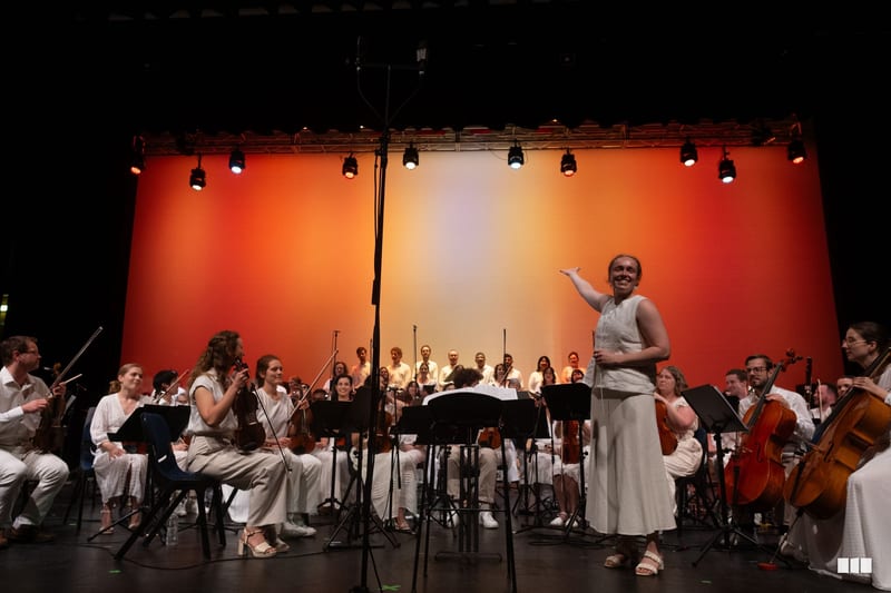 A conductor leads a large orchestra during a performance on stage with warm orange and yellow lighting.
