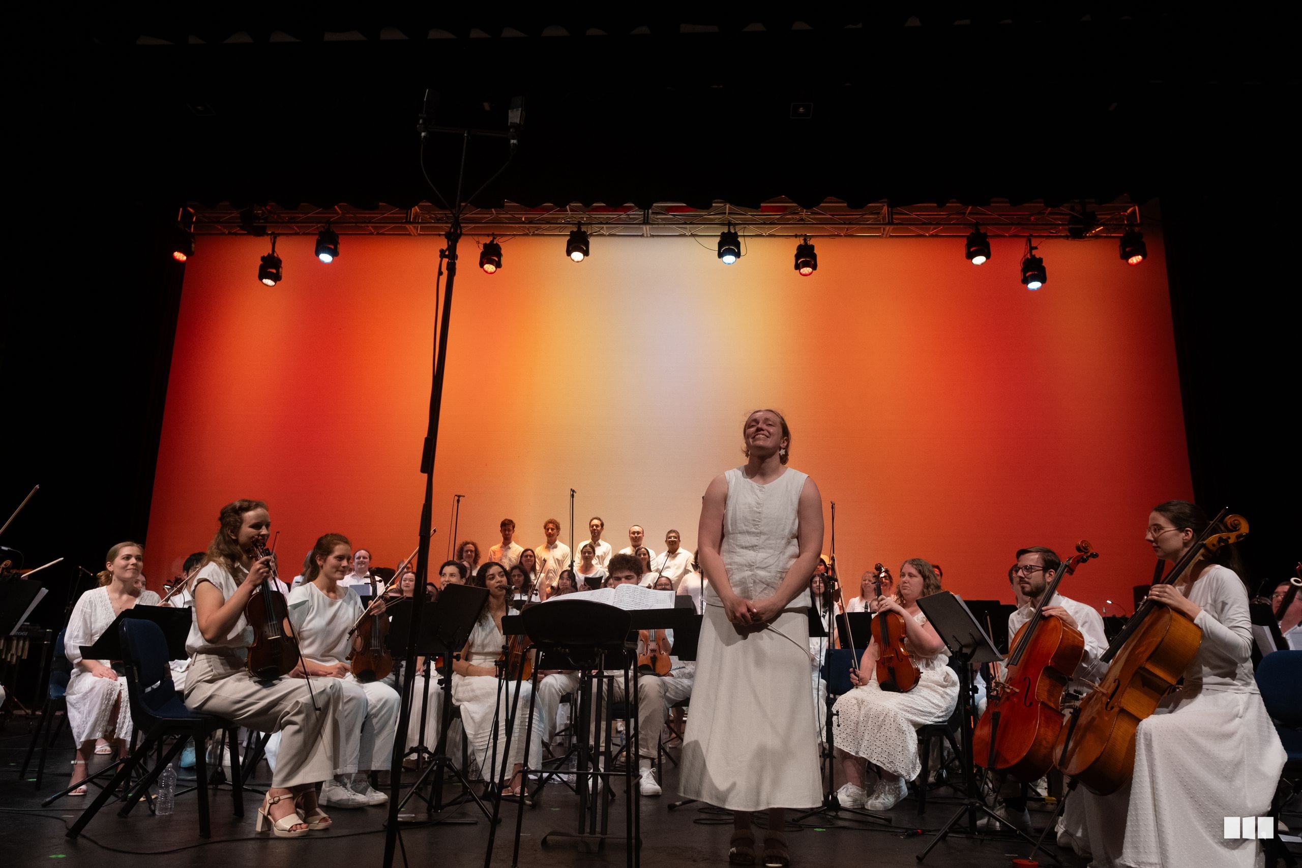 A female soloist in a white dress performs with an orchestra and choir on stage against an orange backdrop.