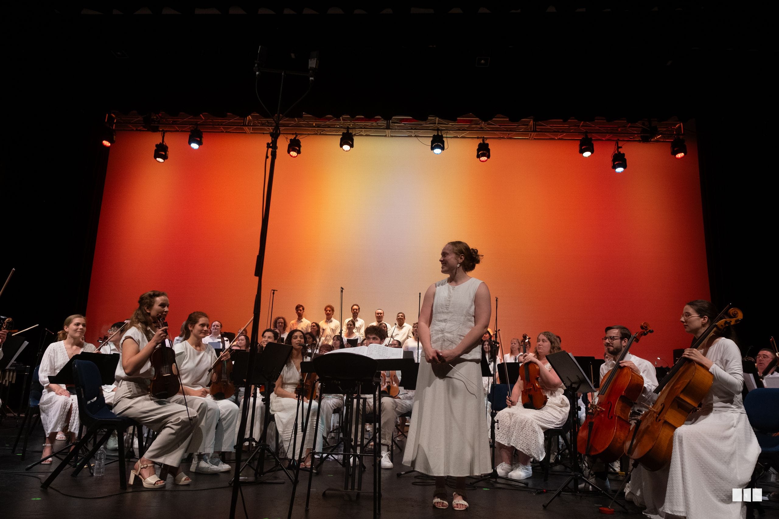 A female soloist in a white dress performs with an orchestra and choir against a vibrant orange backdrop during a concert.