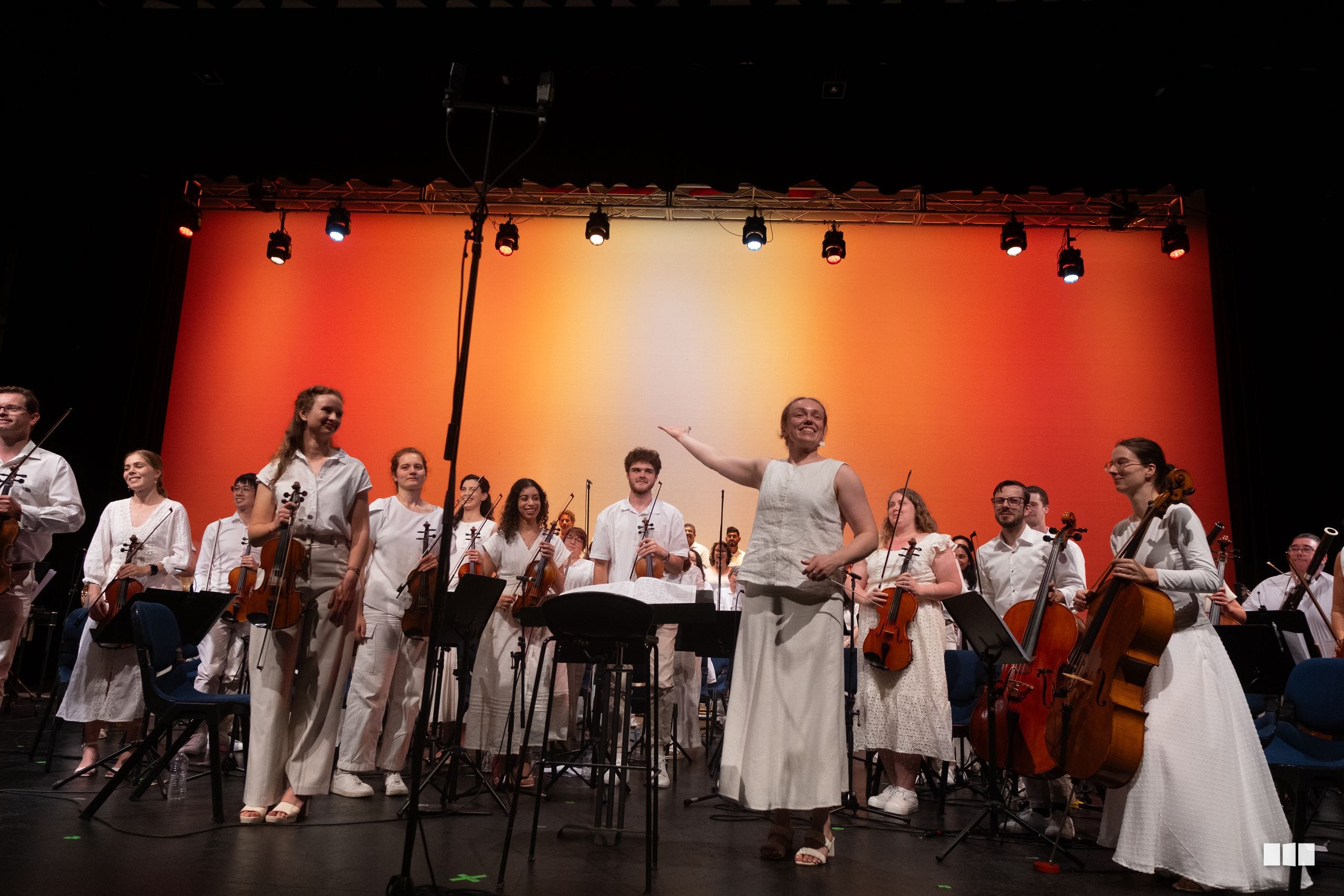 A conductor leads a string orchestra ensemble in white attire during a performance against an illuminated orange backdrop.