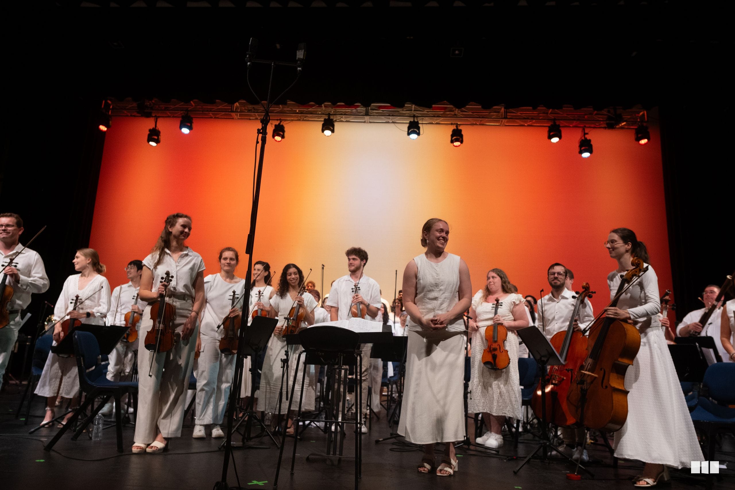 A string orchestra ensemble in white attire takes a bow on stage under warm orange lighting after their performance.