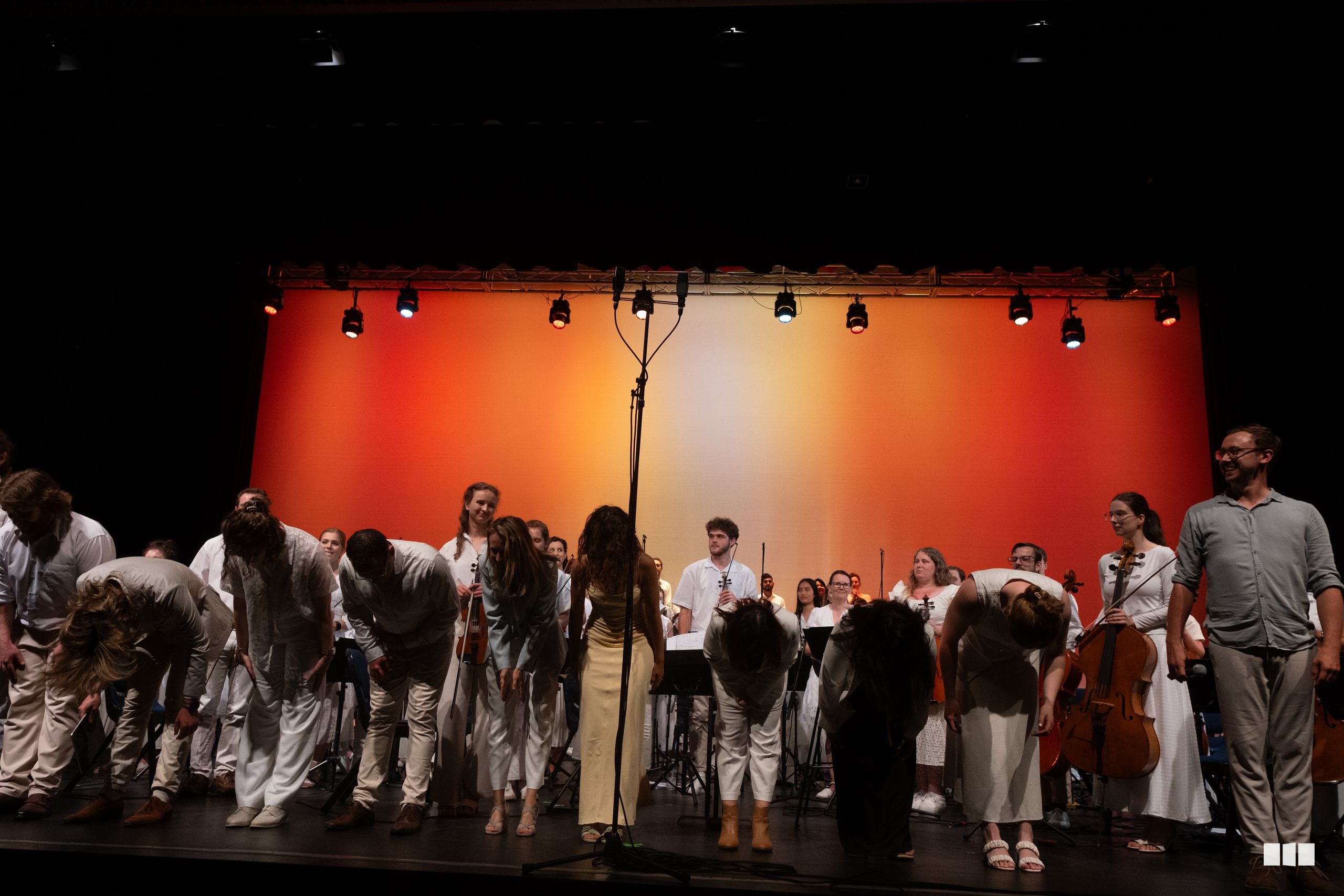 A chamber orchestra and choir ensemble takes a bow on stage under warm orange and yellow stage lighting after a performance.